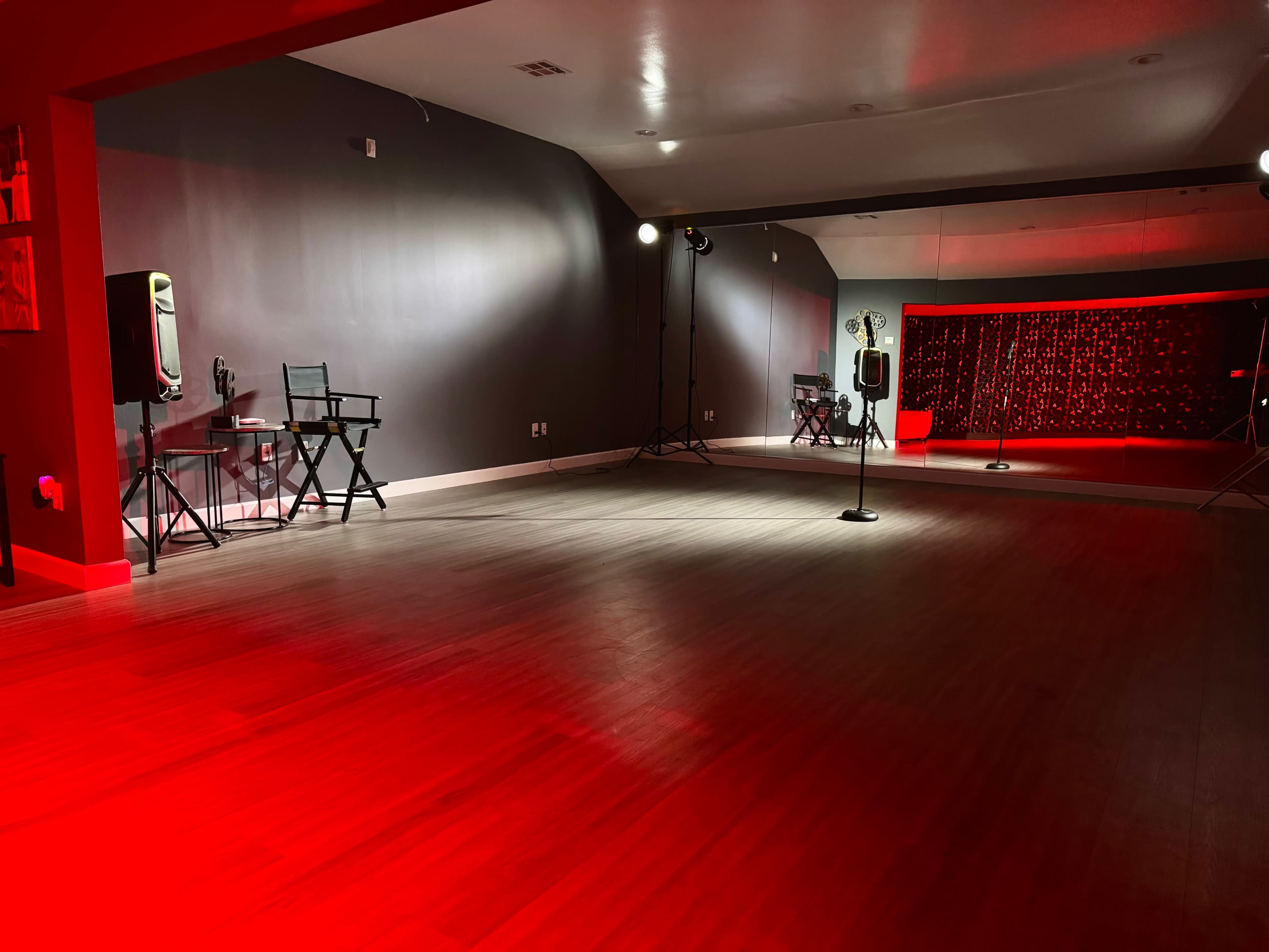 The image shows a sparsely furnished dance studio with a wooden floor, a large mirror, and red lighting effects.