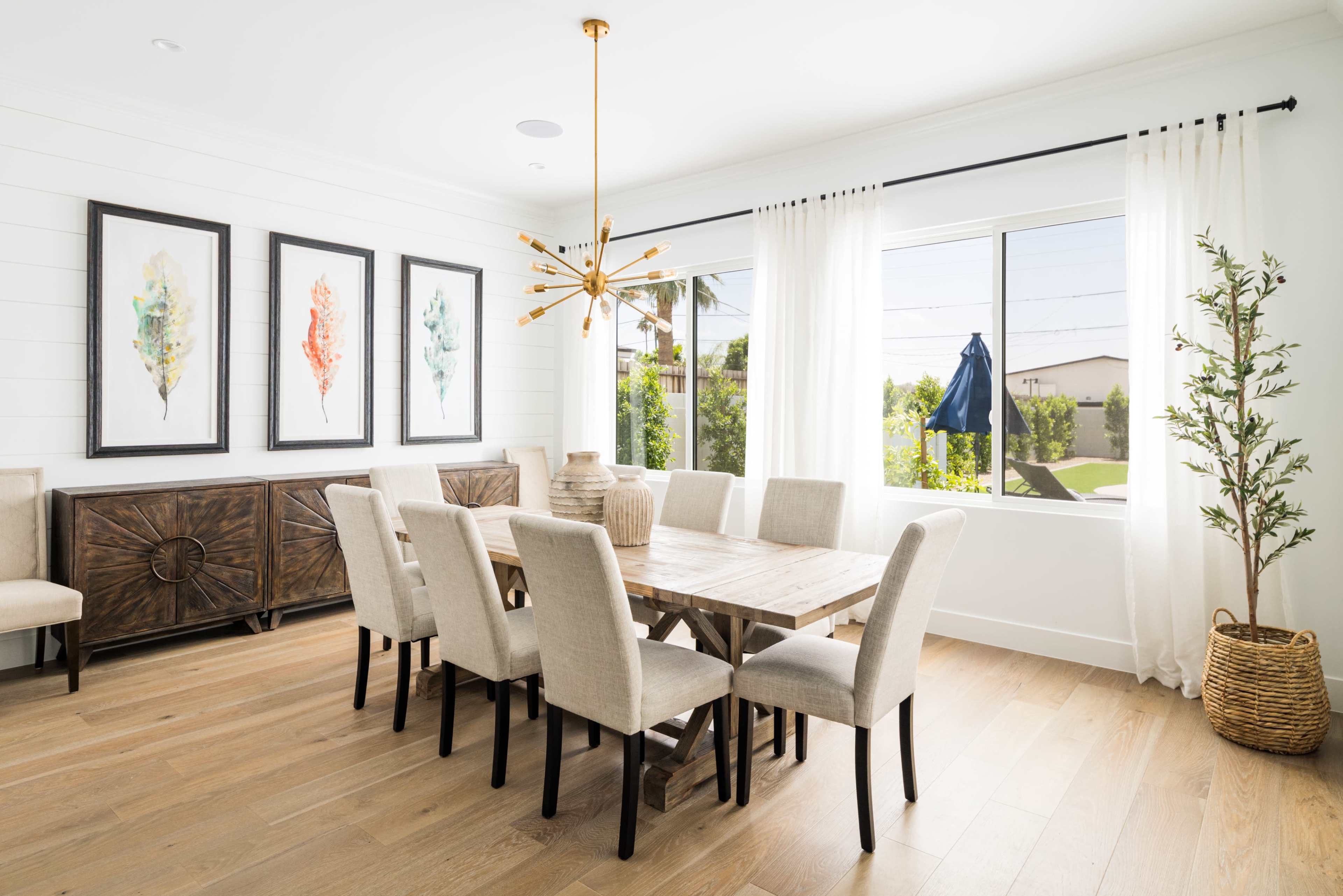 A modern dining room with a wooden table surrounded by beige chairs, framed artwork on the walls, and large windows letting in natural light.