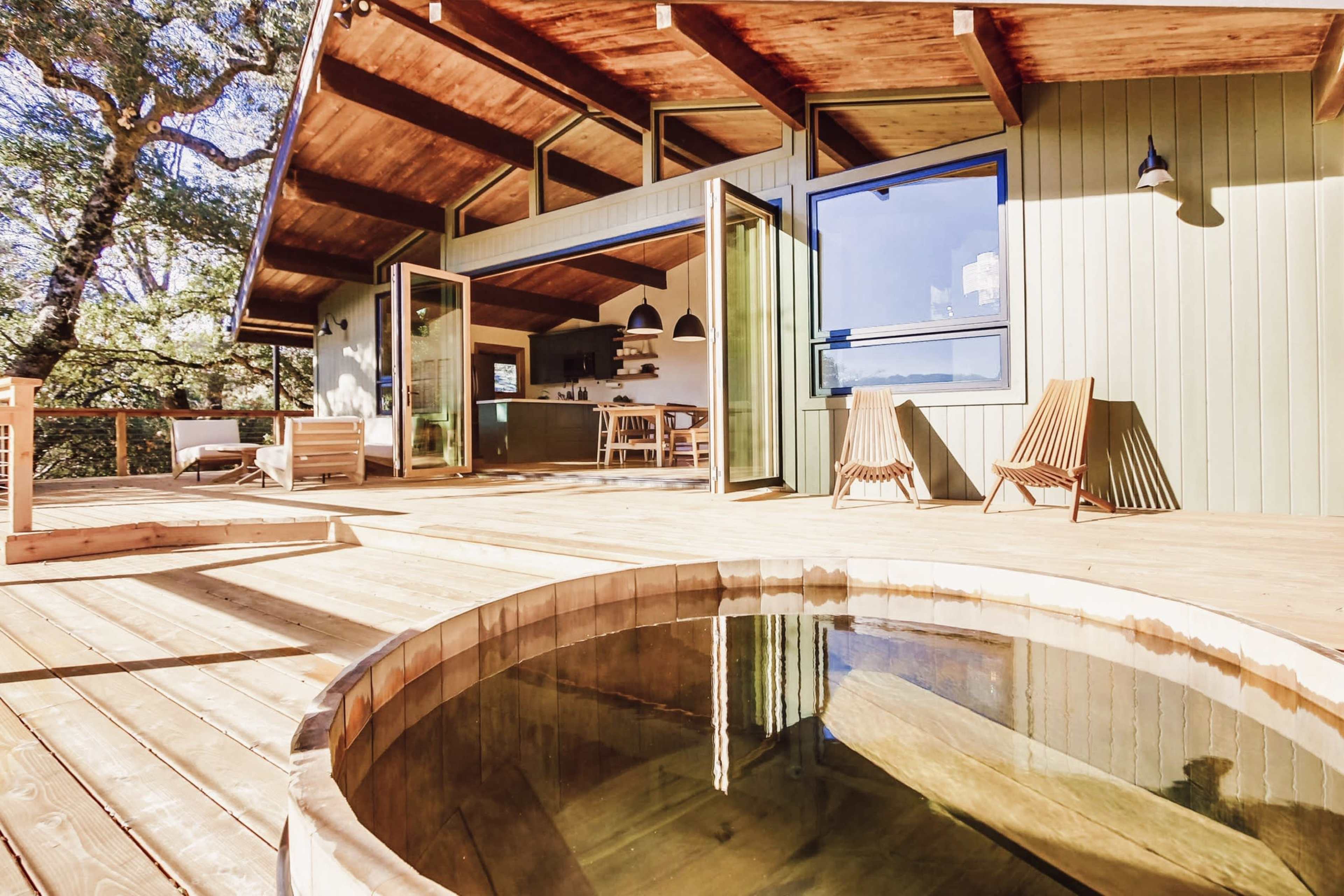 A wooden deck features an outdoor hot tub with two wooden lounge chairs and expansive views of the surrounding trees.