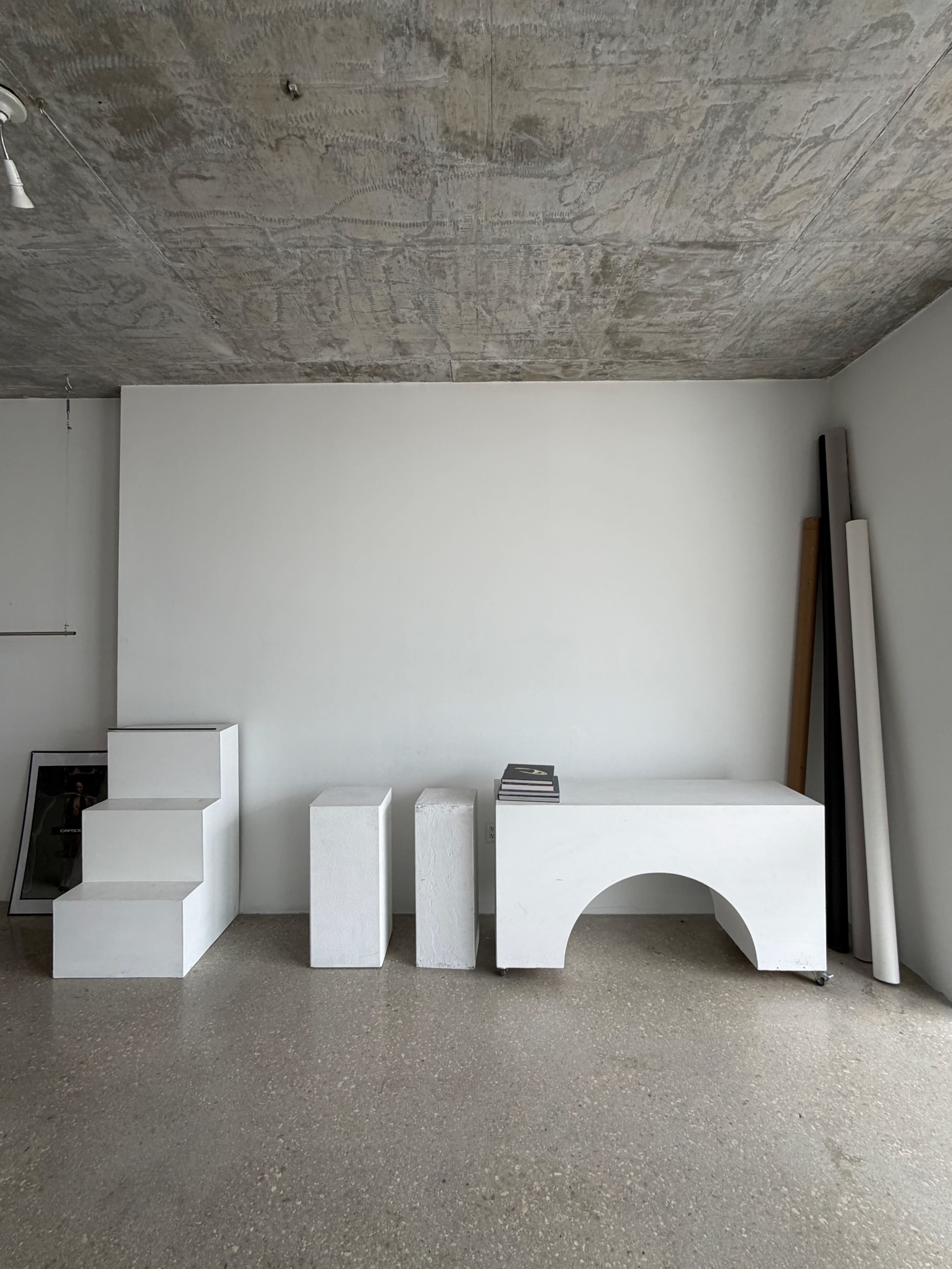 The image shows a minimalist interior featuring several white geometric furniture pieces, including a stair-like structure and cylindrical forms, against a plain wall.