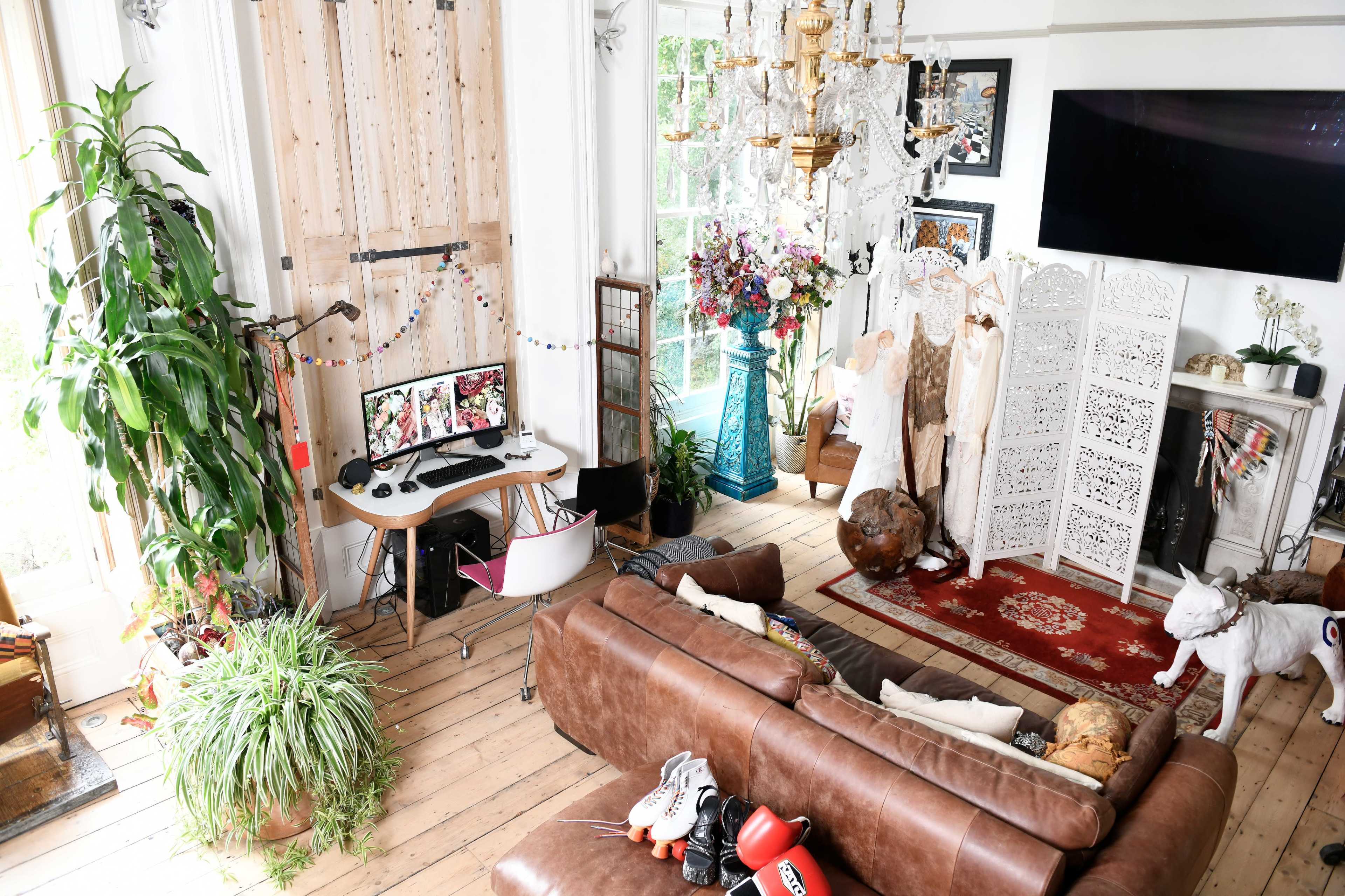 The image shows a bright, eclectic living room with a brown leather couch, a computer setup, a large chandelier, decorative plants, and a variety of artworks and furnishings.