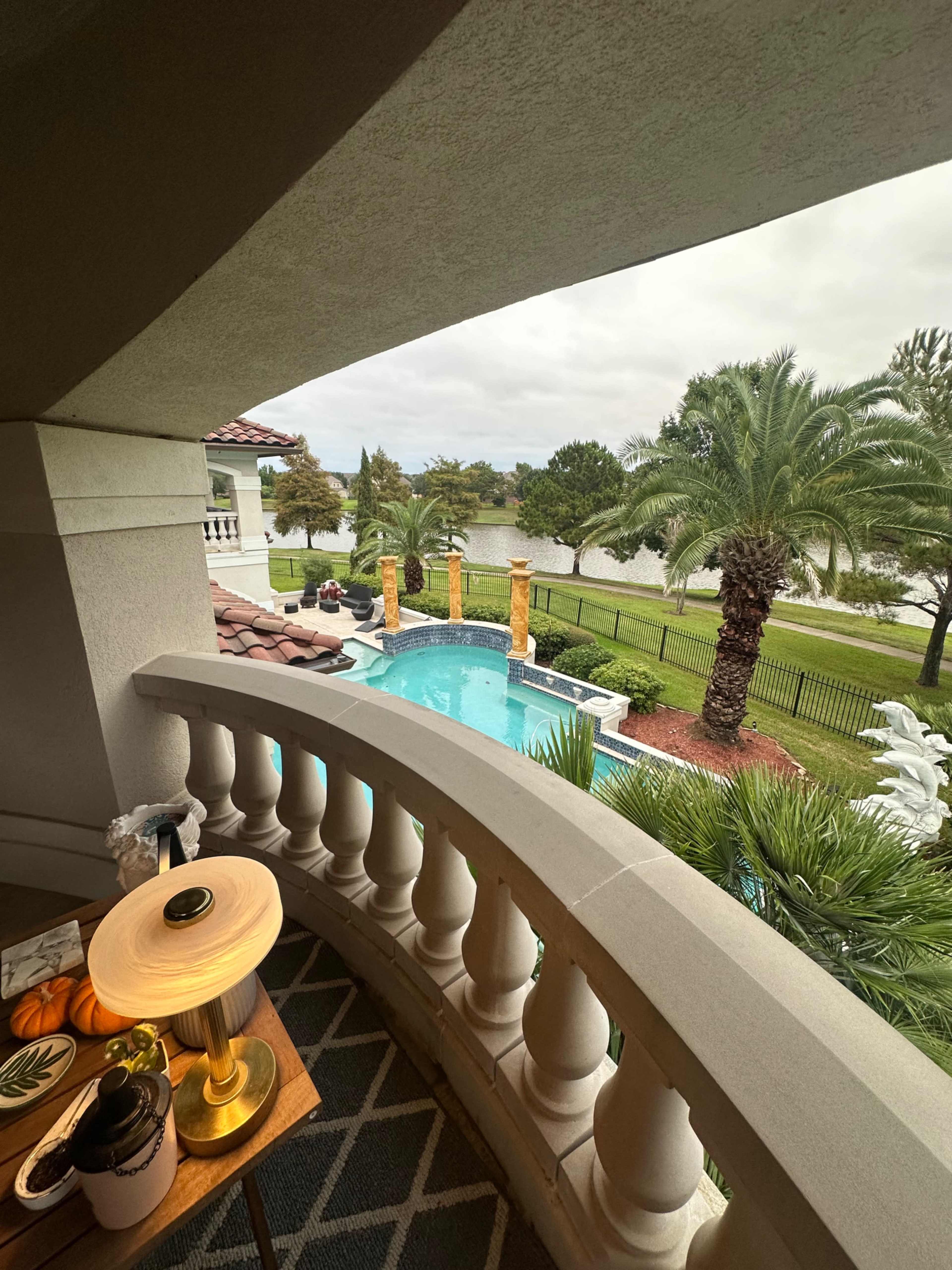The image shows a balcony view overlooking a pool area with palm trees and a body of water in the background.