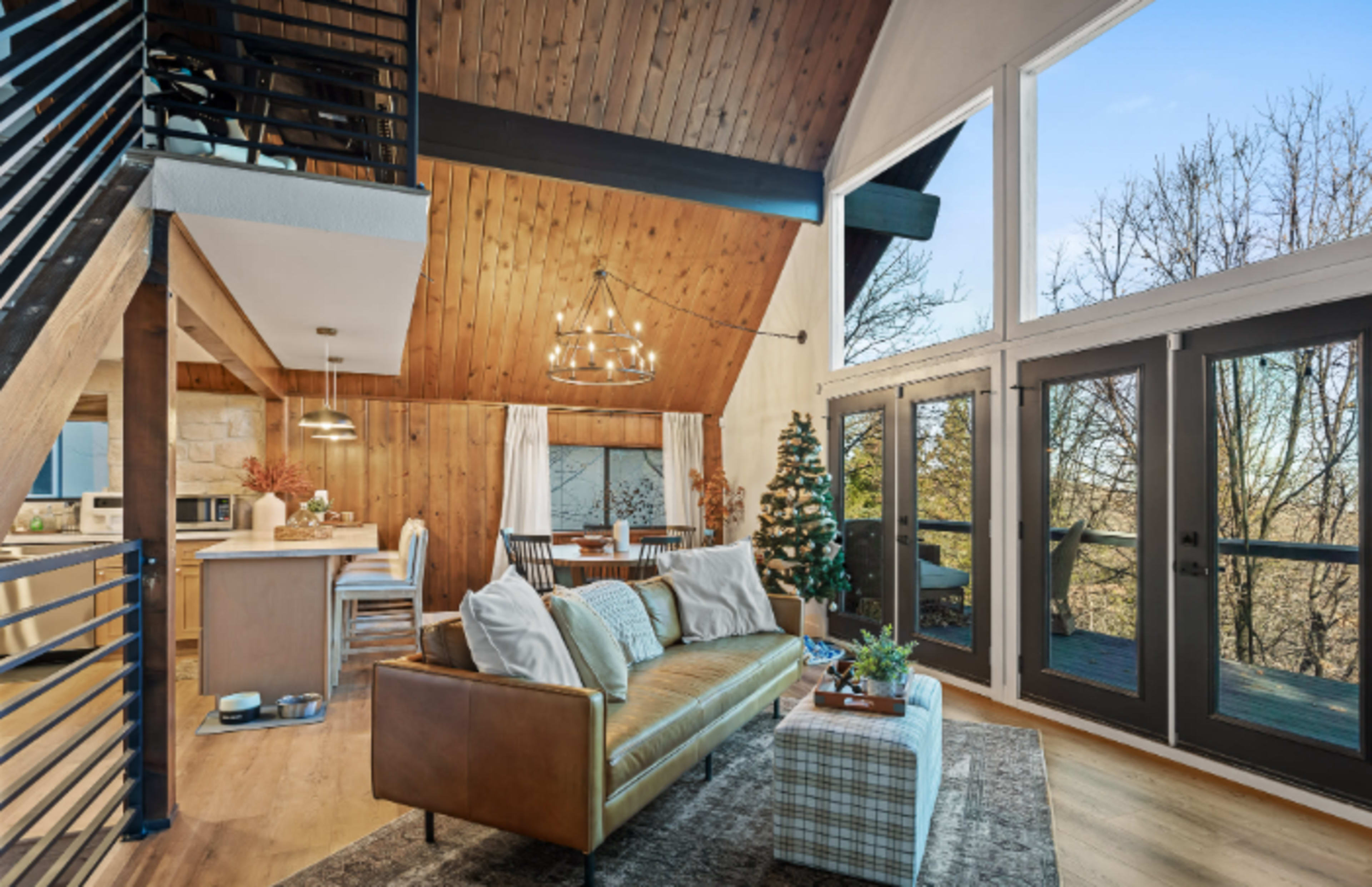 The image shows a modern living area with a wooden ceiling, large windows, and a staircase leading to a loft, adorned with a Christmas tree and cozy furniture.