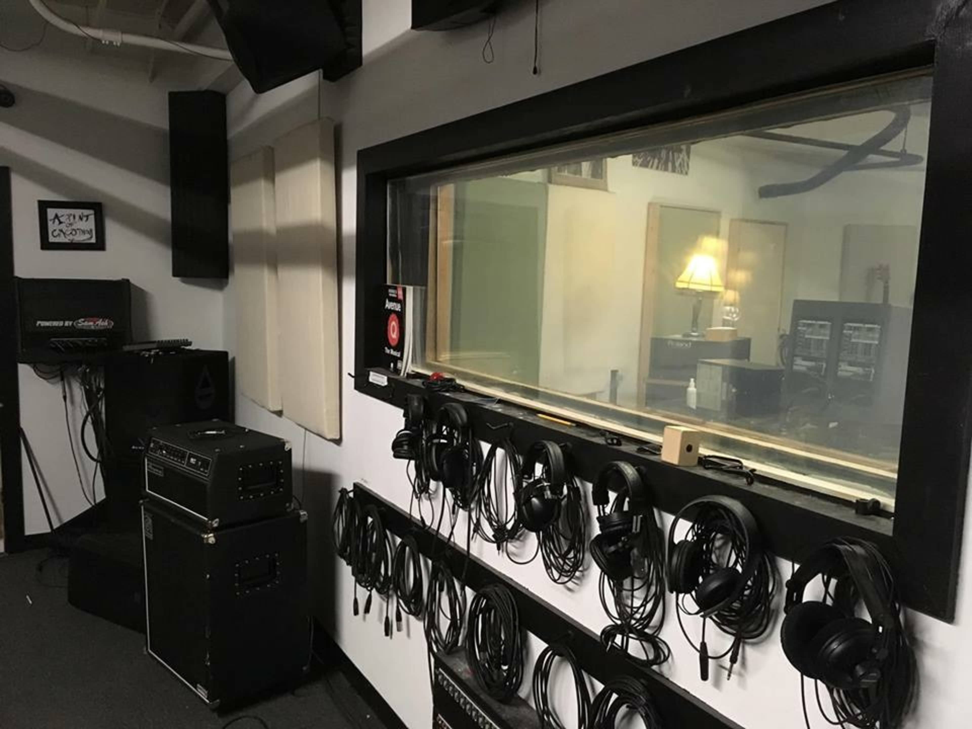The image shows a recording studio with a control room featuring a large glass window, headphones hanging on a wall, and audio equipment in the corner.