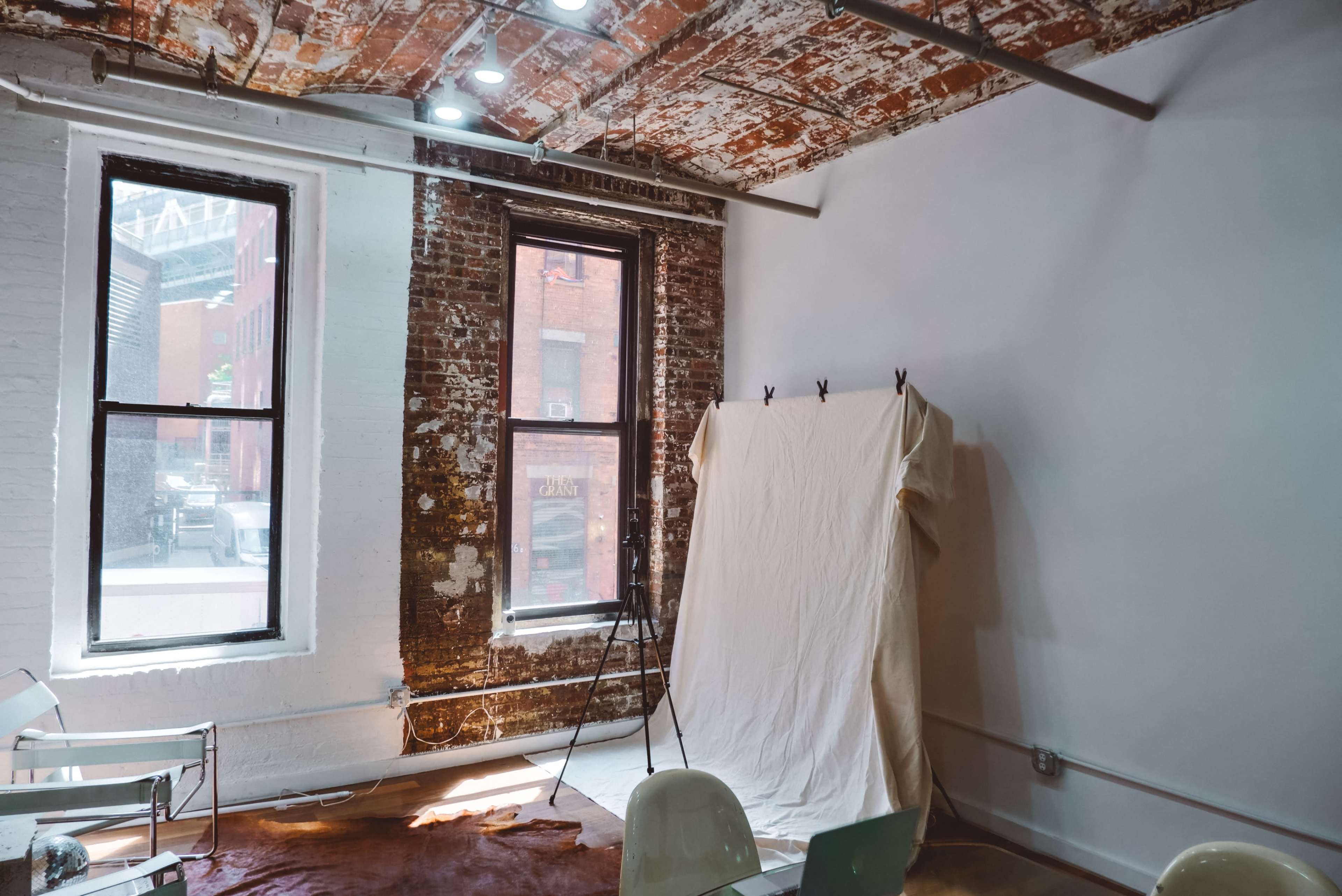 A minimalist room with exposed brick walls, large windows, and a draped backdrop on a stand.