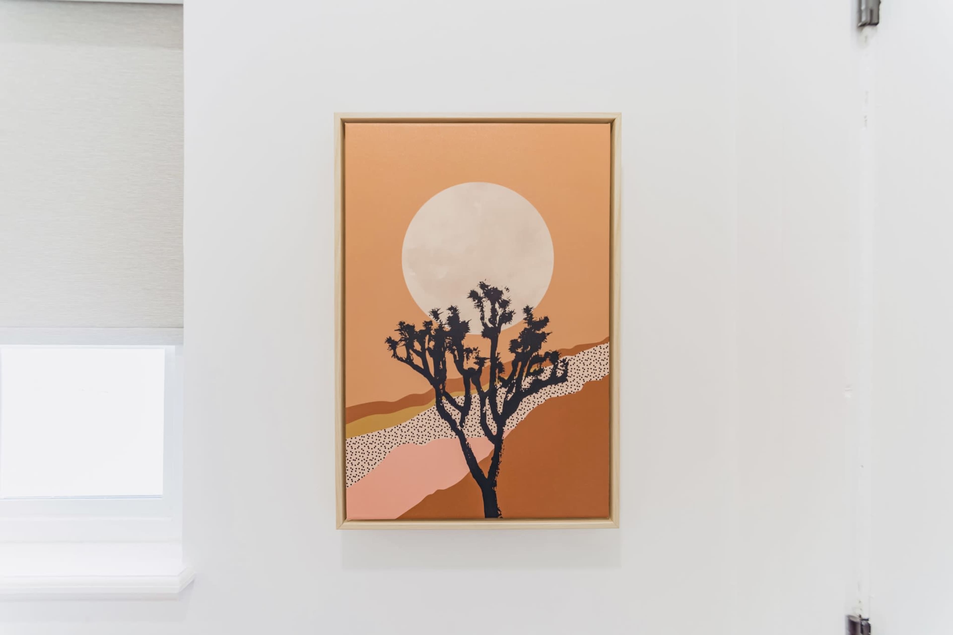 A framed artwork depicting a silhouette of a tree against a large sun and abstract landscape in warm tones is hung on a white wall next to a window.