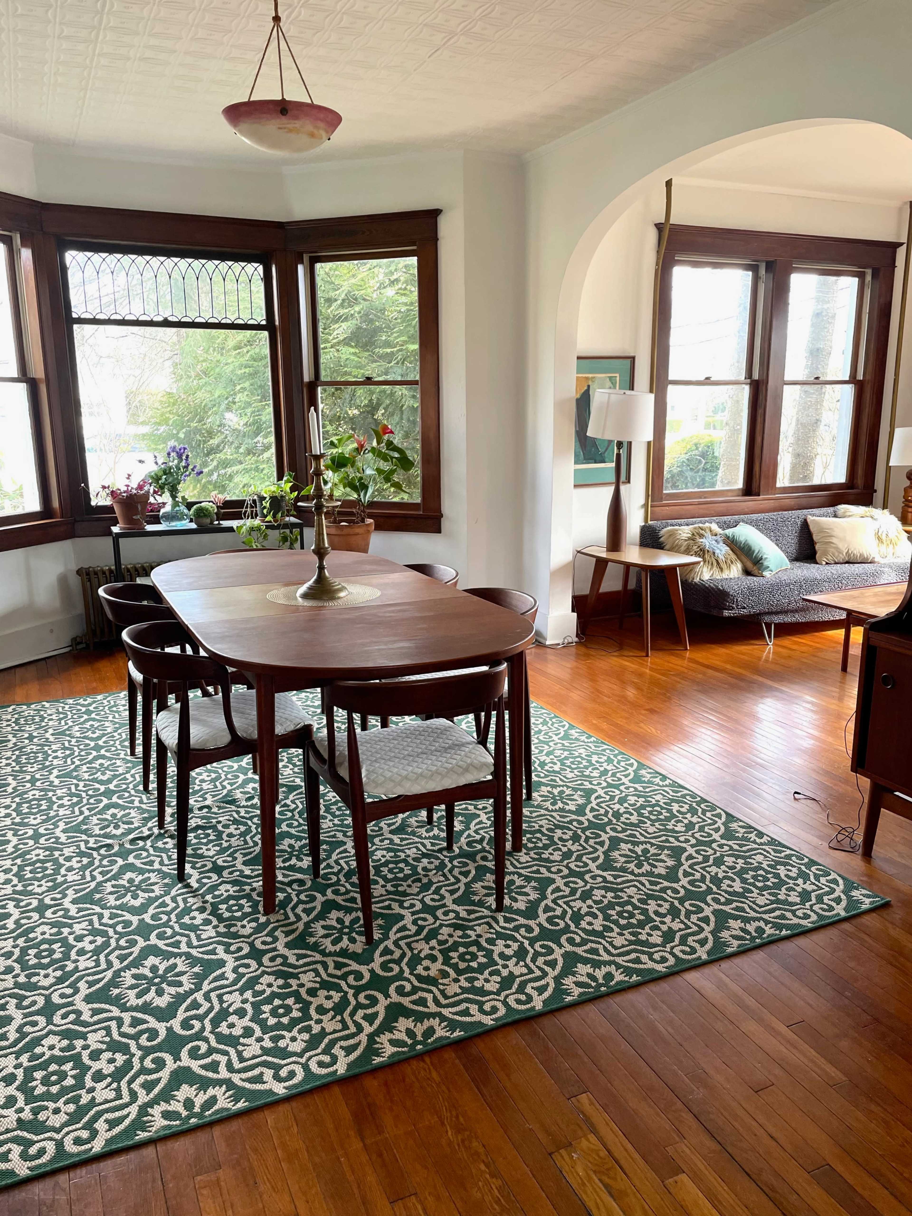 A dining area features a wooden table surrounded by chairs, a patterned rug, and large windows with plants beside a sofa in a well-lit room.