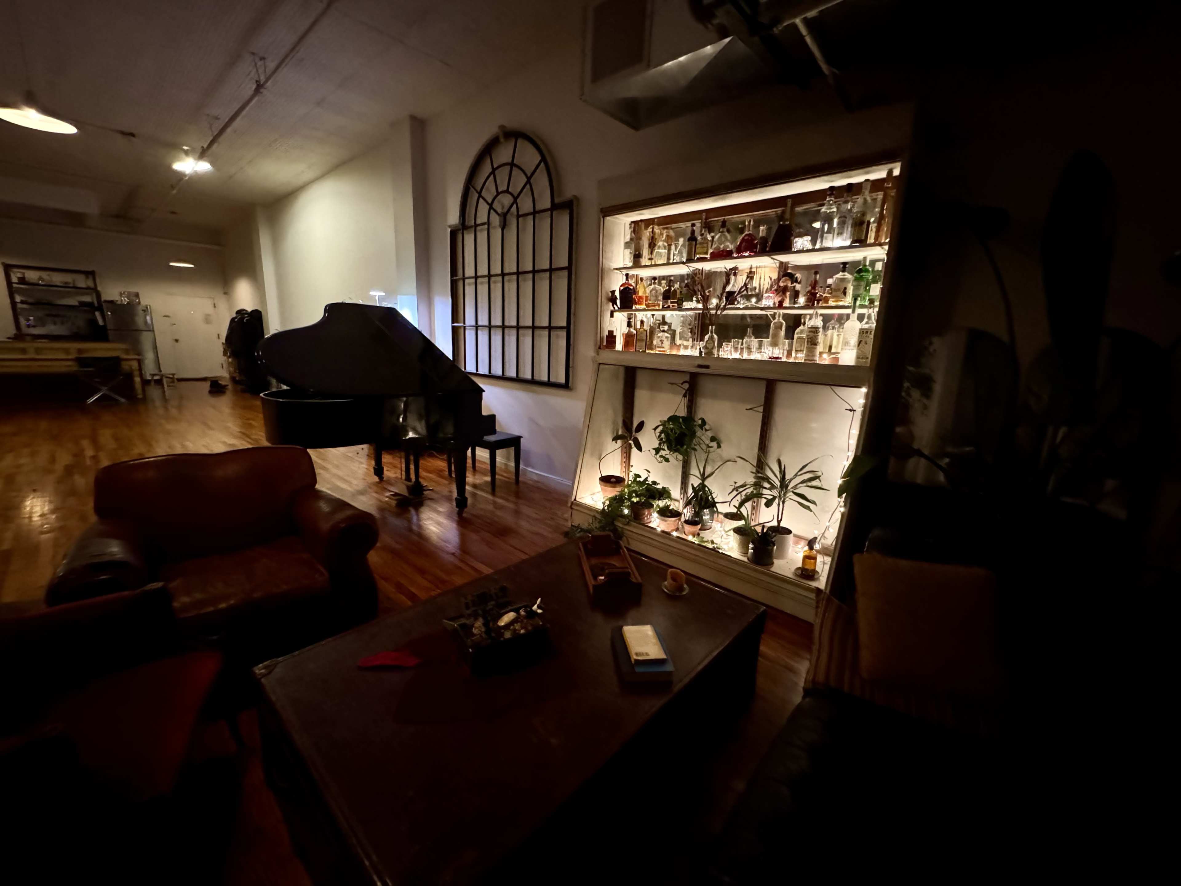 The image depicts a dimly lit room with a grand piano, a coffee table, and a shelf displaying various glass bottles and plants.