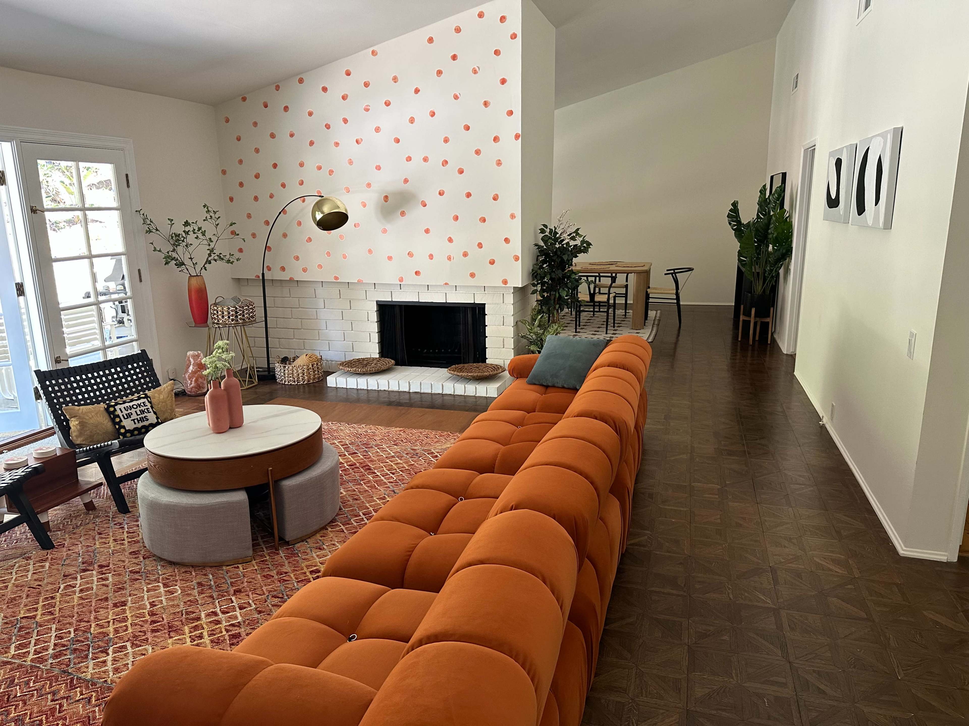 The image shows a spacious living area featuring an orange sofa, a round coffee table, a fireplace, and decorative plants, with a dining area visible in the background.