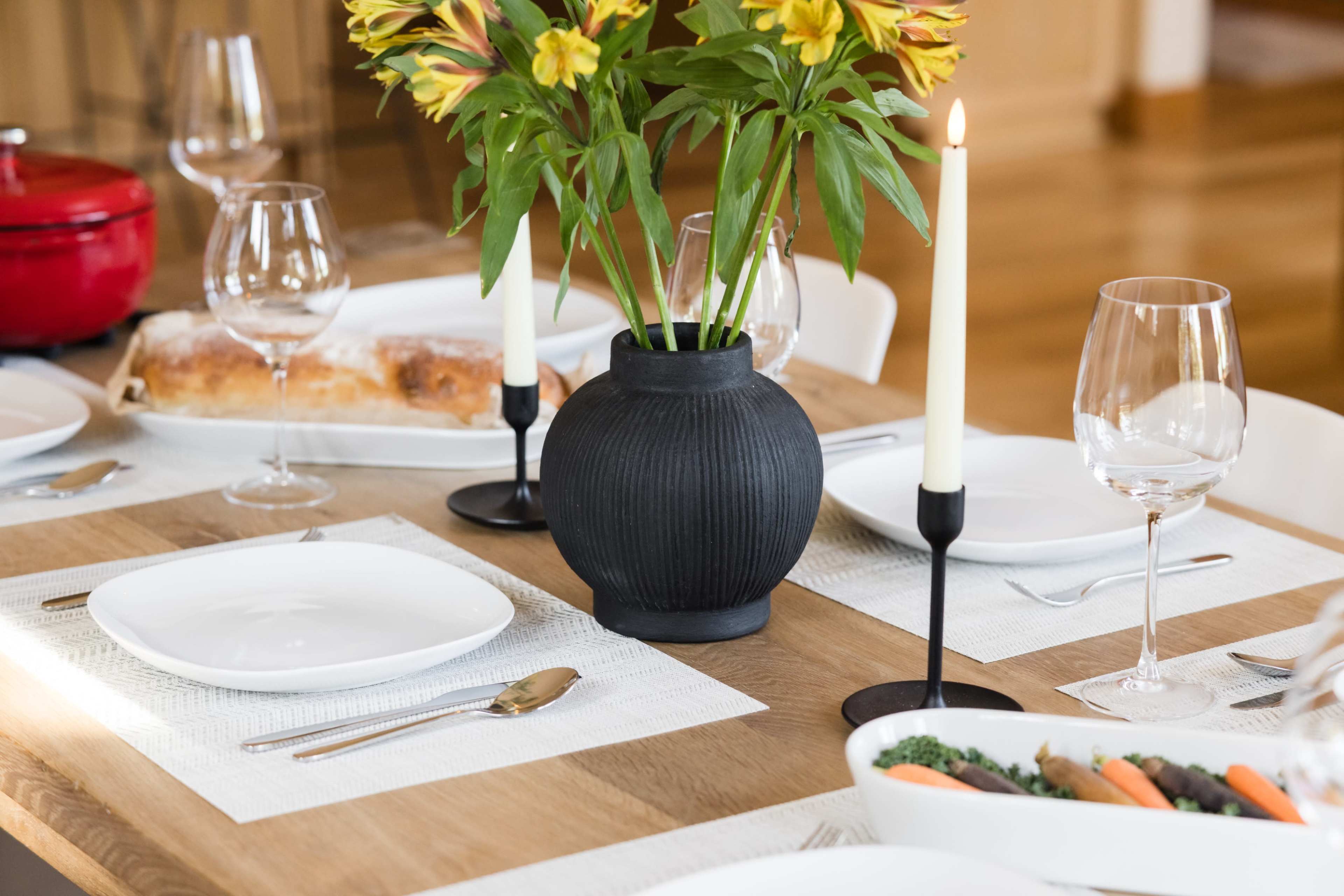 A dining table is set with white plates, wine glasses, a candle, and a vase of flowers, accompanied by a bread dish and a bowl of vegetables.