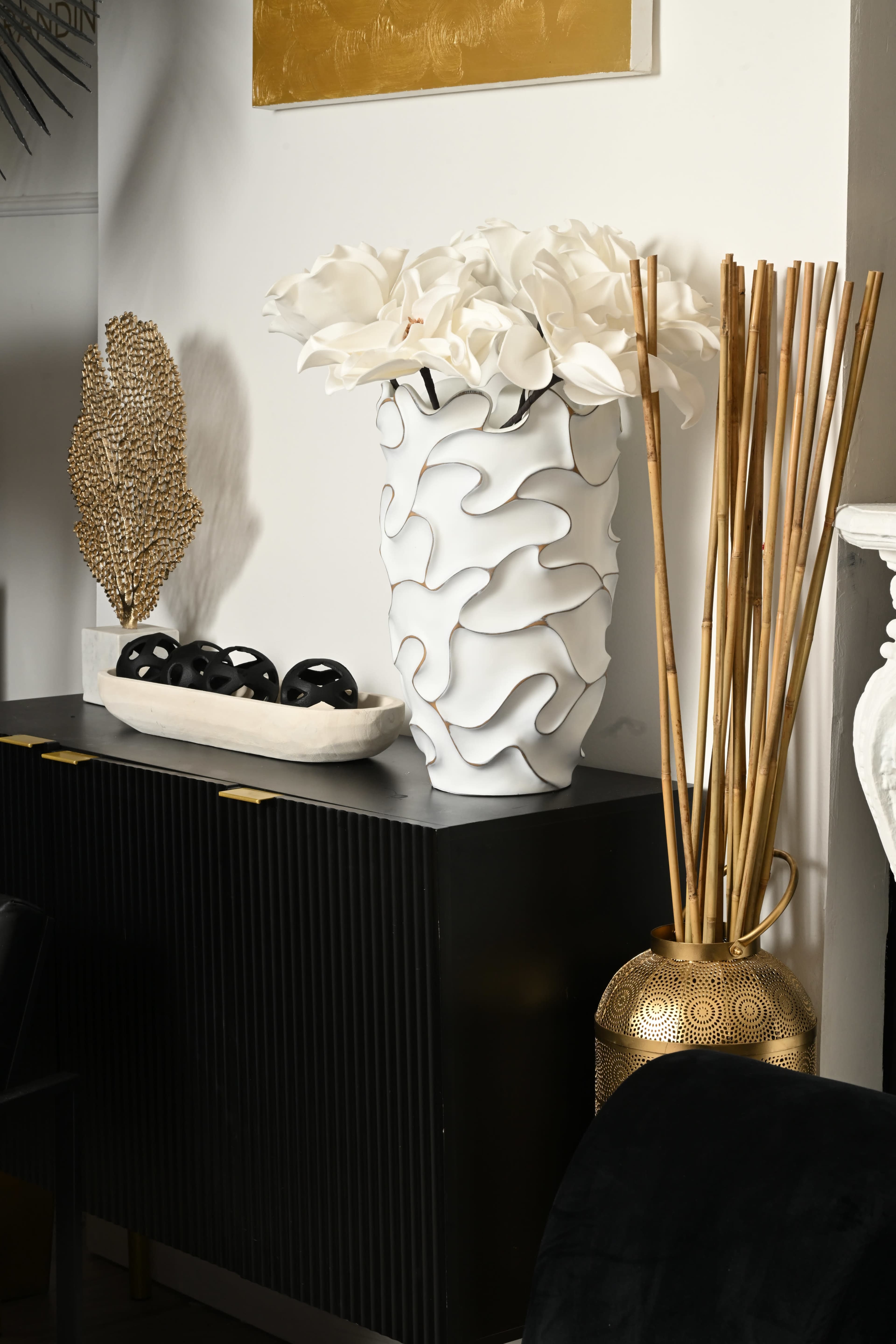 A black cabinet displays a white wavy vase filled with artificial white flowers, alongside decorative elements including a gold leaf sculpture, a bowl of black spheres, bamboo sticks, and a gold metal accent piece.