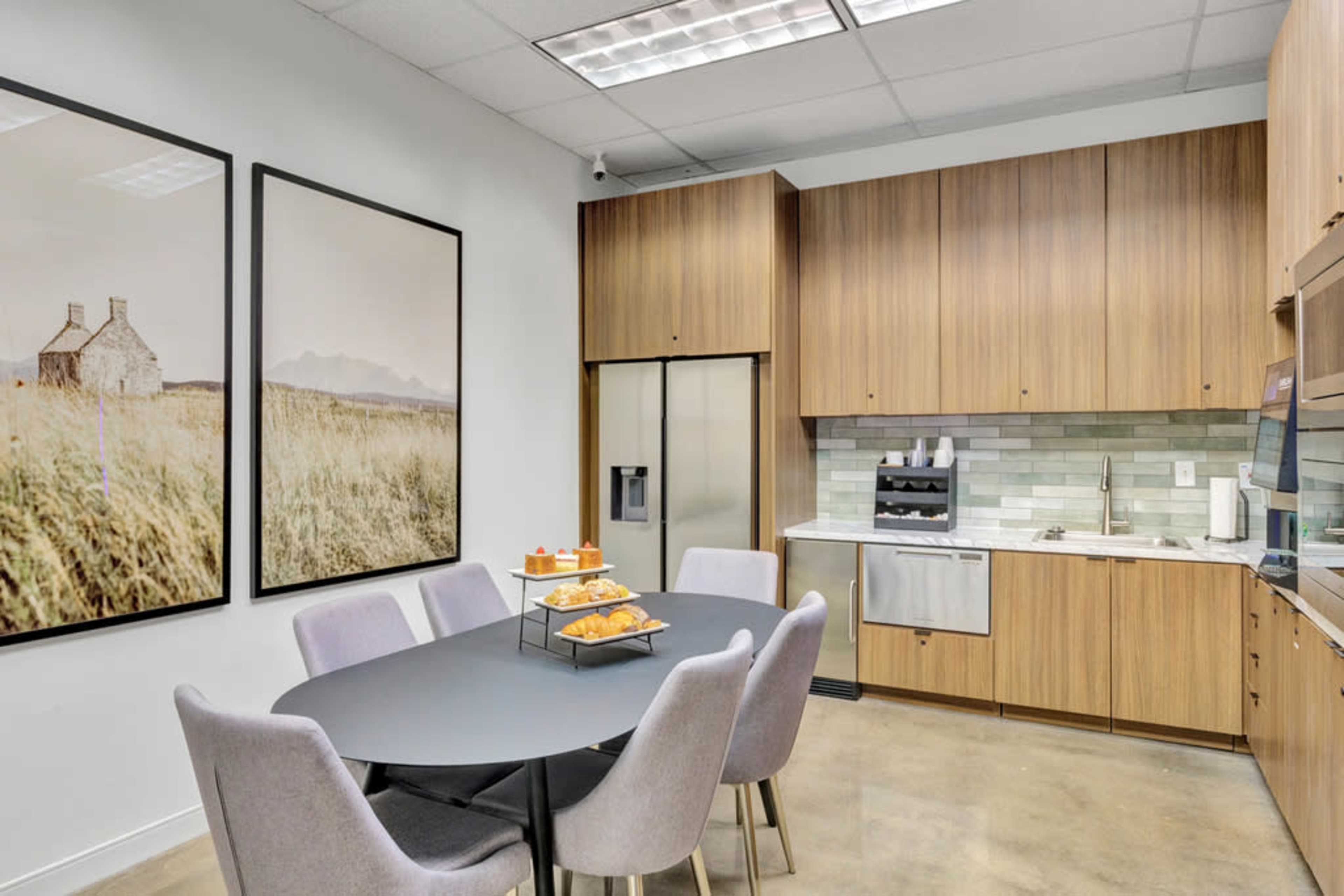 The image shows a modern kitchen area featuring wood cabinetry, a round table with gray chairs, and framed artwork on the walls.