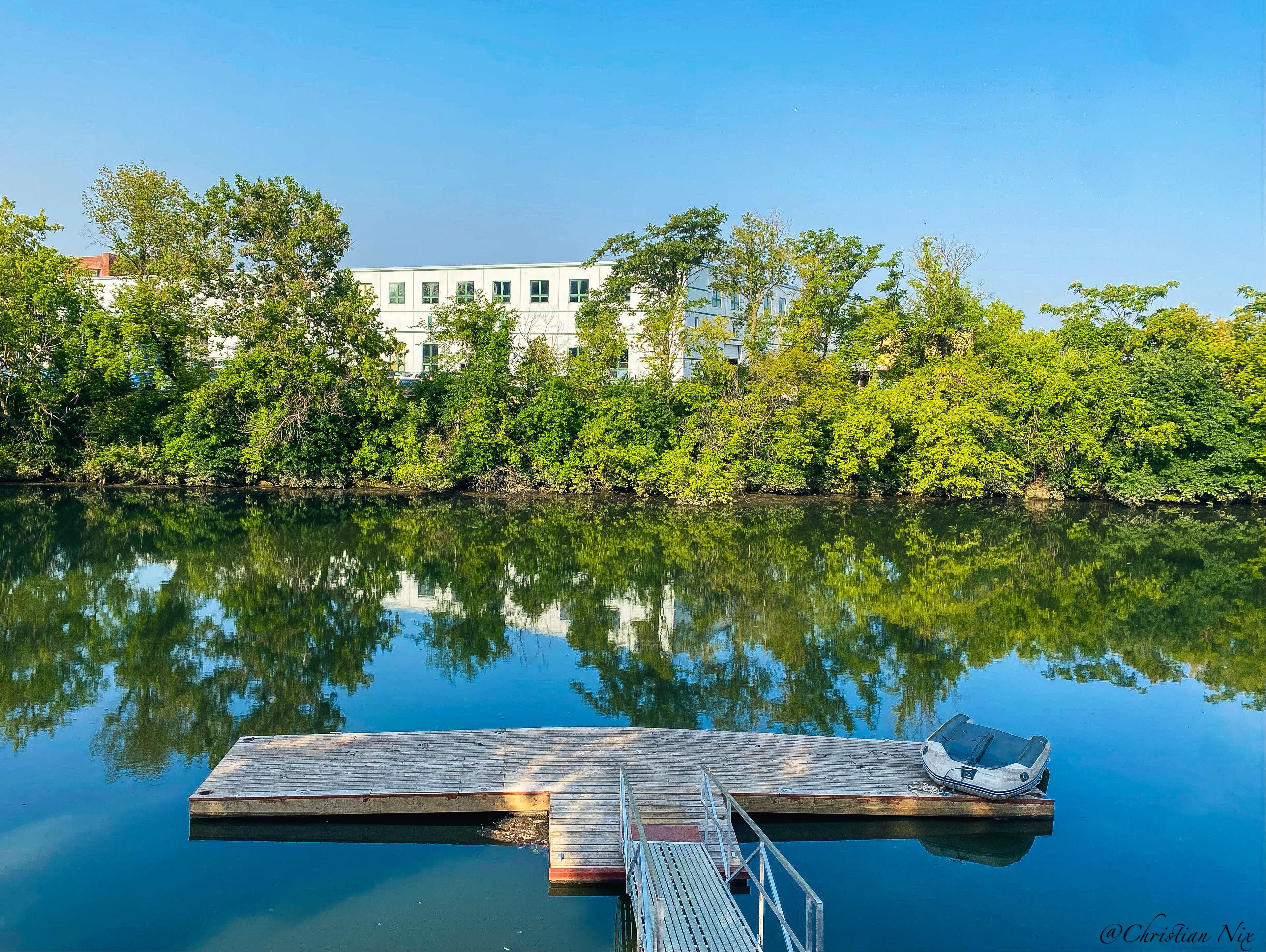 A wooden dock extends over a calm river, reflecting trees and a building on the opposite bank.