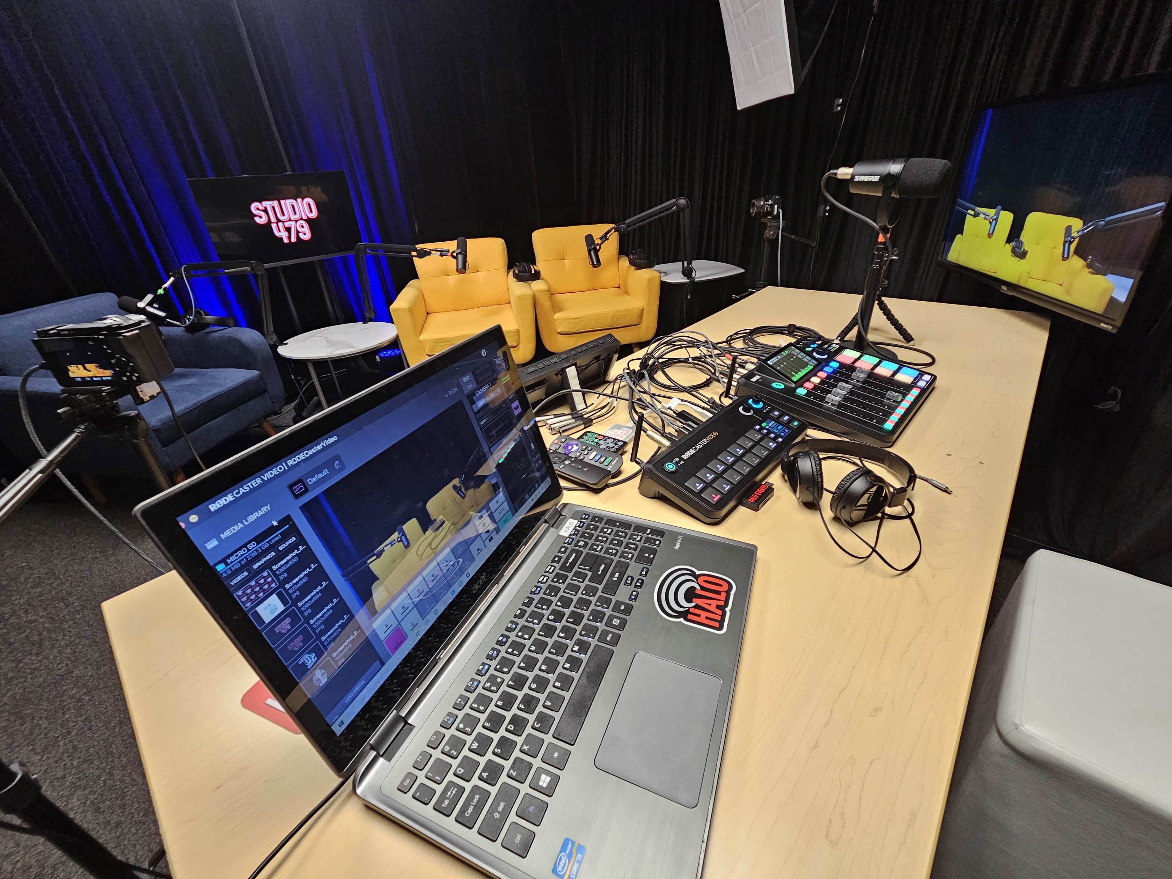 A multimedia studio setup features a laptop, audio equipment, microphones, and two yellow chairs against a backdrop with studio lighting.