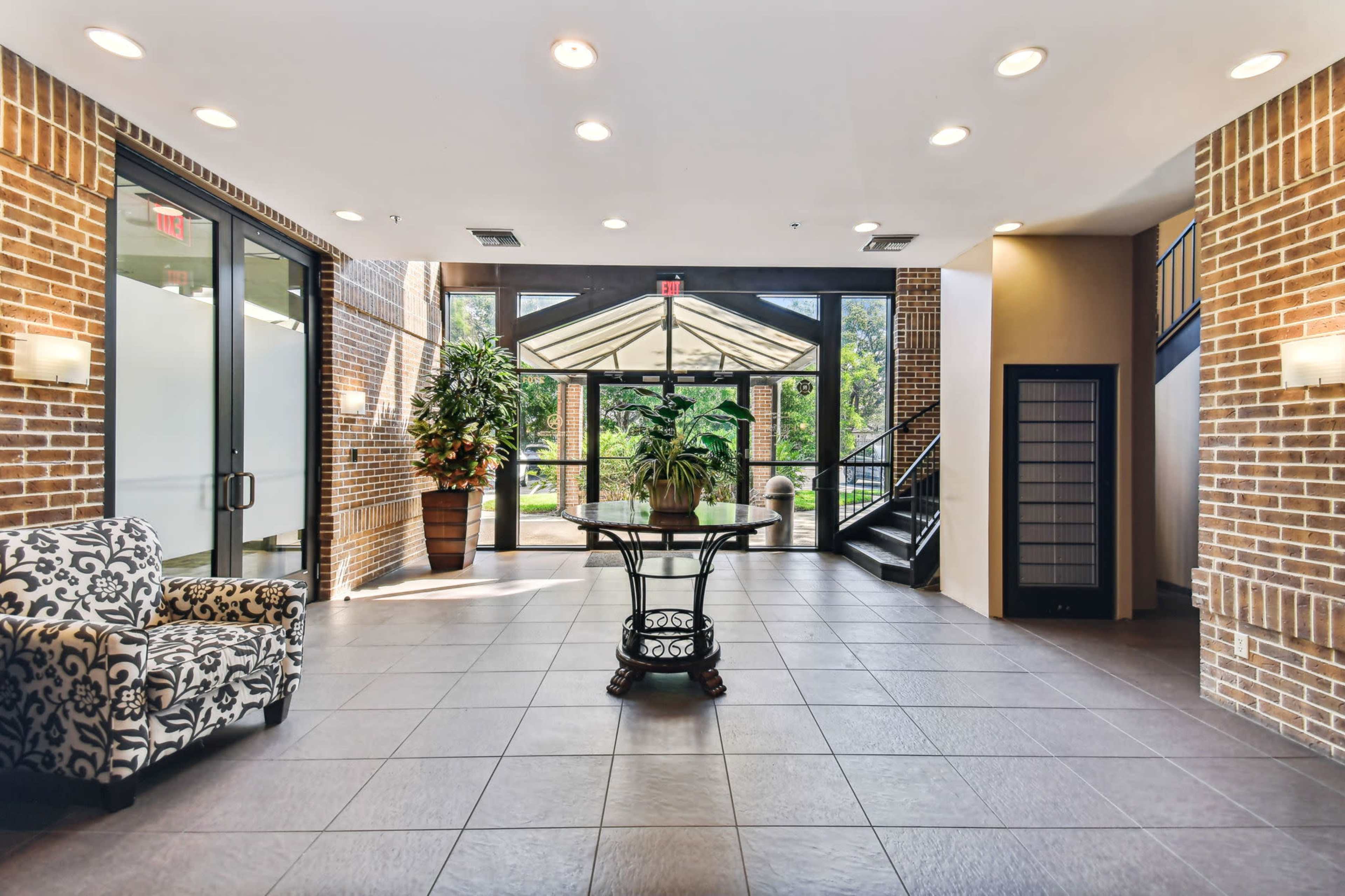 The image shows a modern lobby with a patterned chair, potted plants, and large glass doors leading outside.