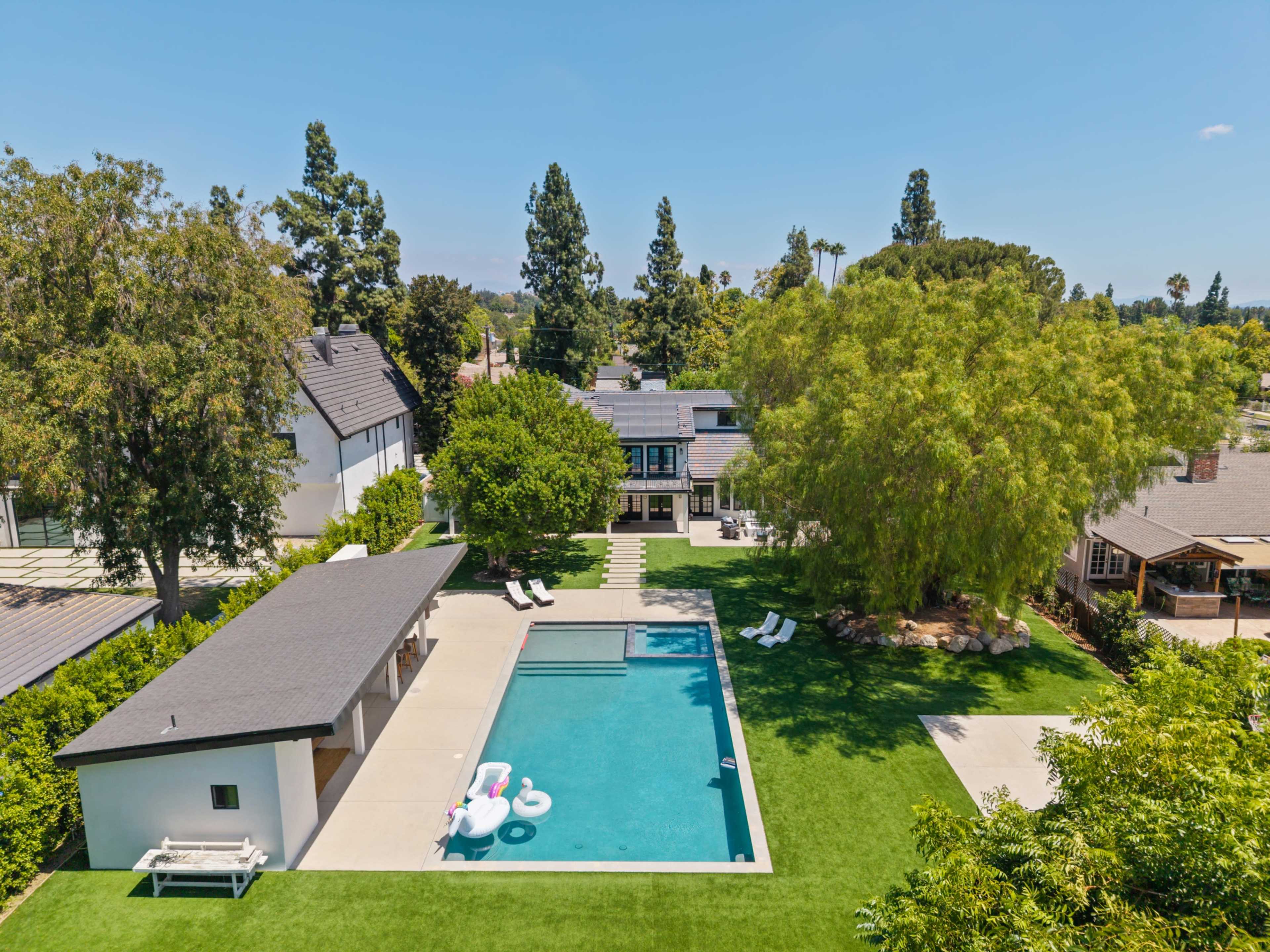 The image shows a spacious backyard with a rectangular swimming pool surrounded by green grass, lounge chairs, and a nearby structure, all set against a clear blue sky.