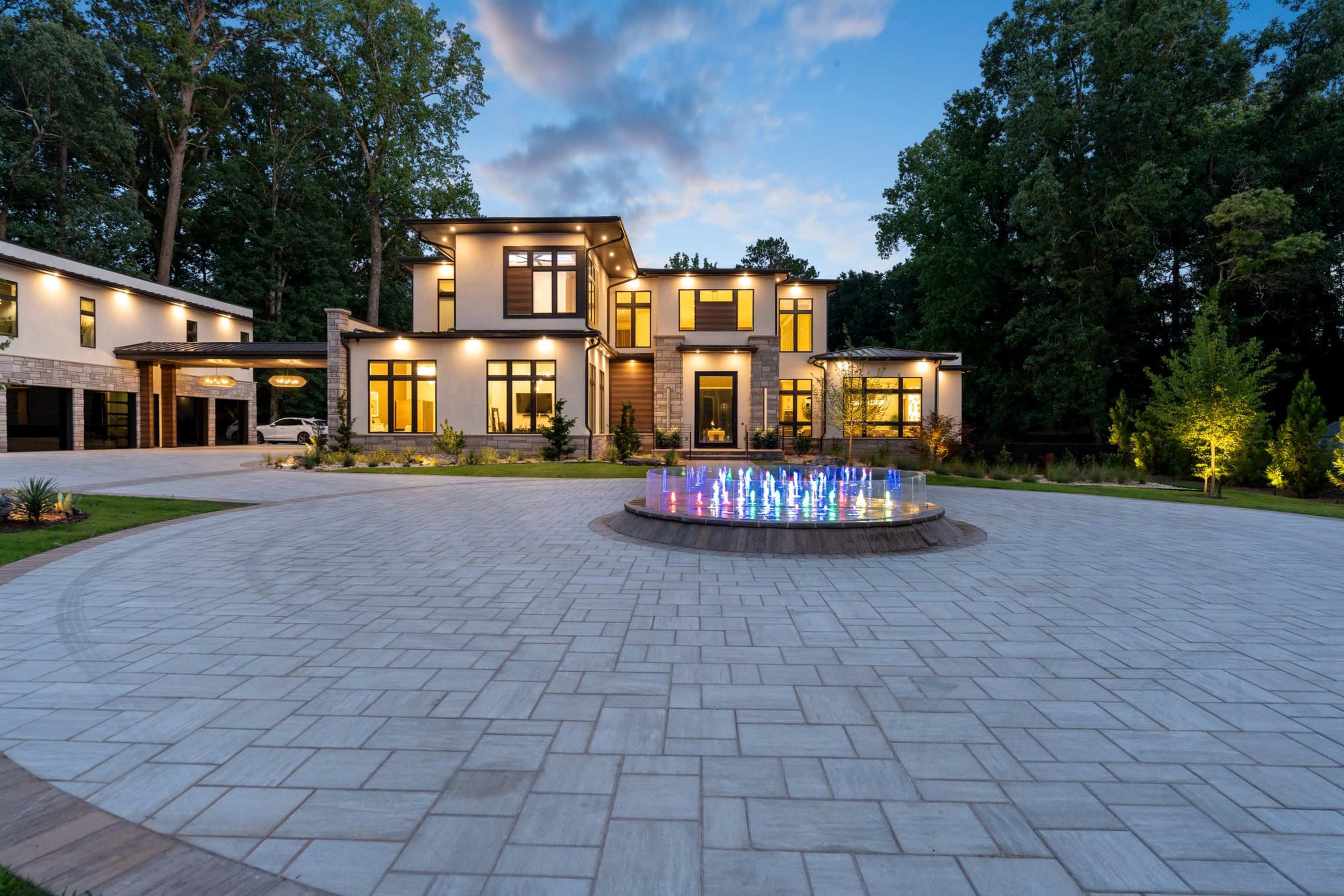 A modern two-story house with large windows is set beside a circular driveway featuring a vibrant illuminated fountain.