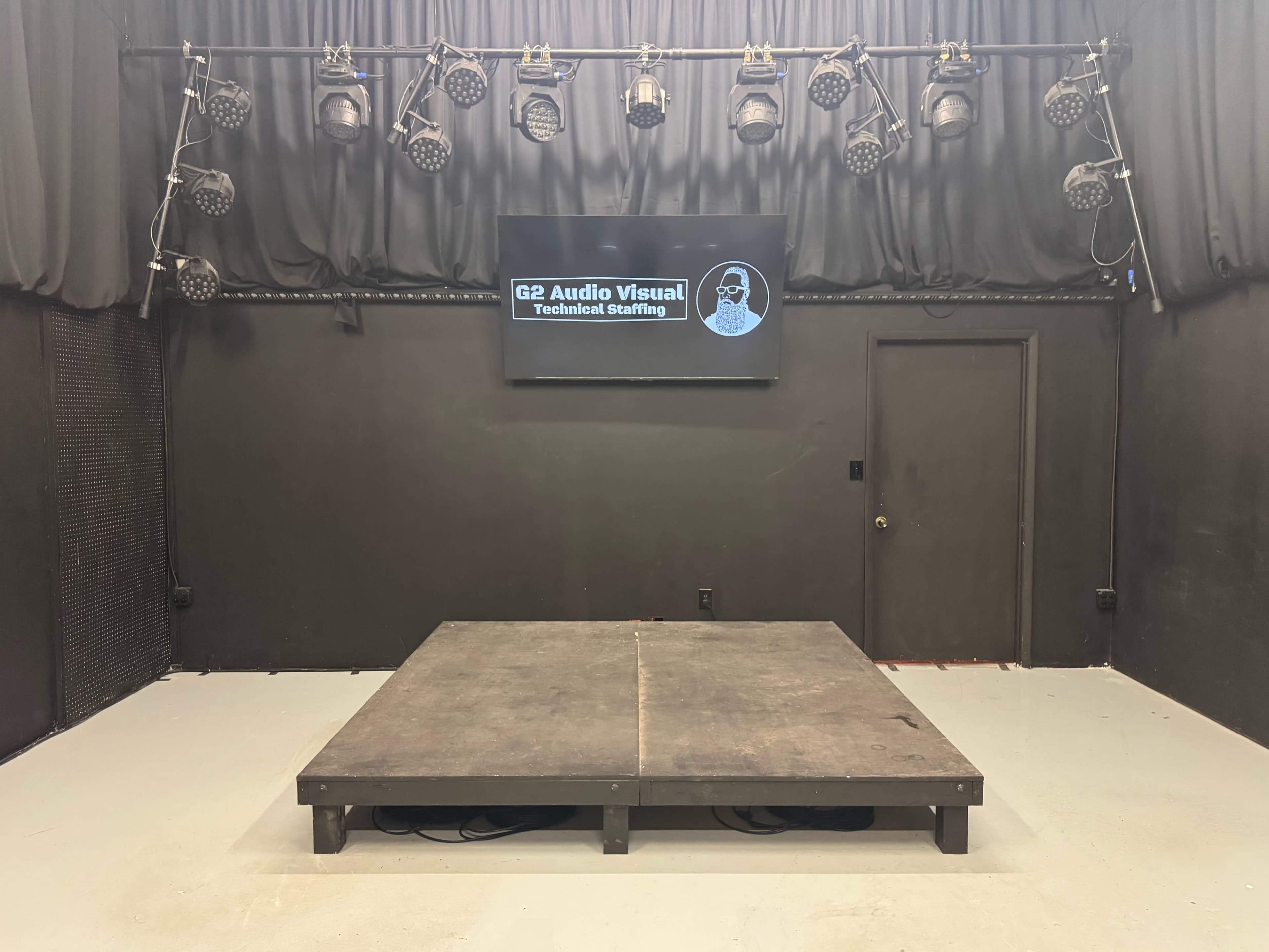 The image shows a small performance stage with a black backdrop, equipped with several overhead lights and a monitor displaying the logo of G2 Audio Visual Technical Staffing.