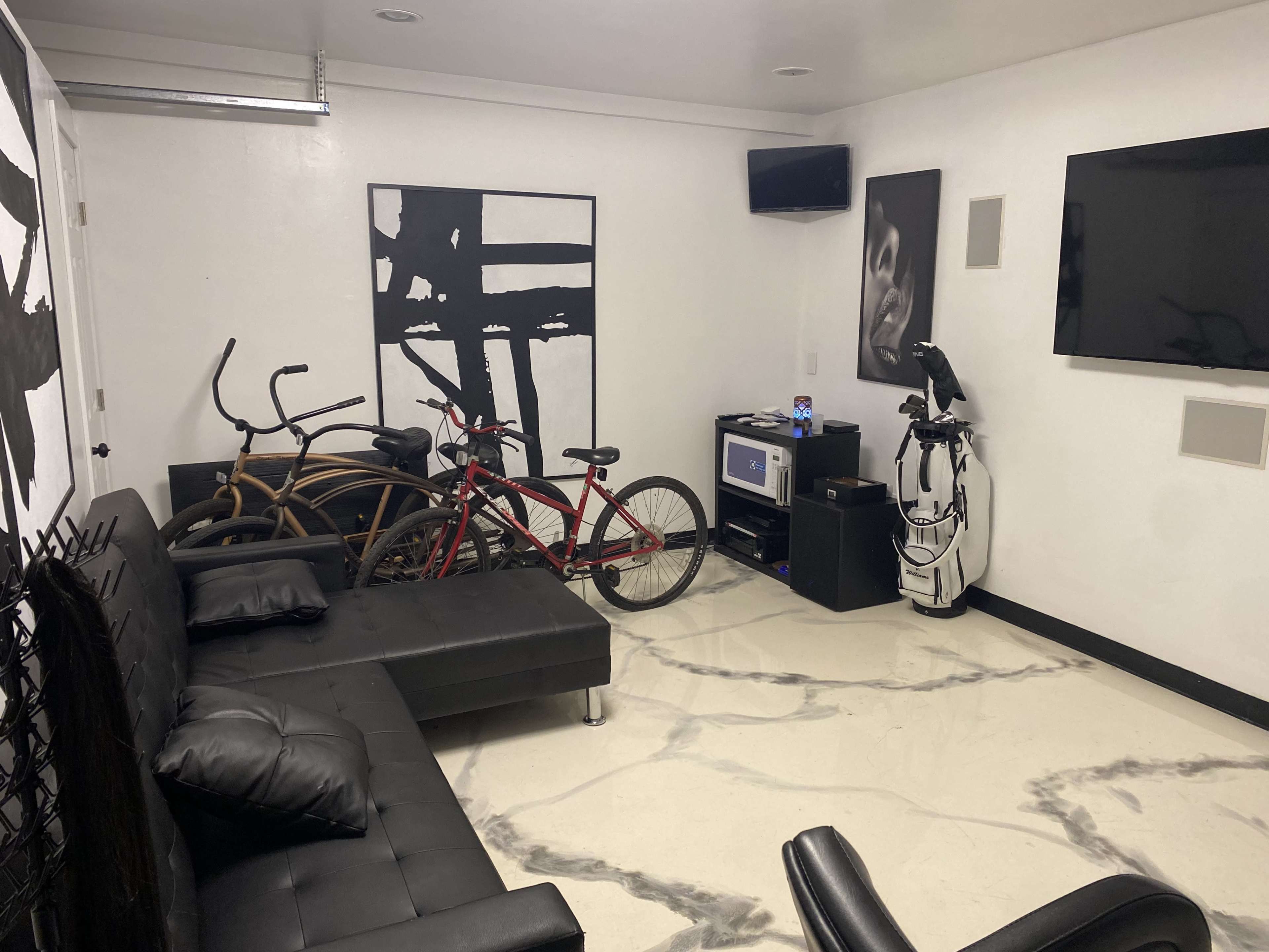 A modern room features two bicycles parked near a sleek black couch, a television on the wall, and minimalist artwork.