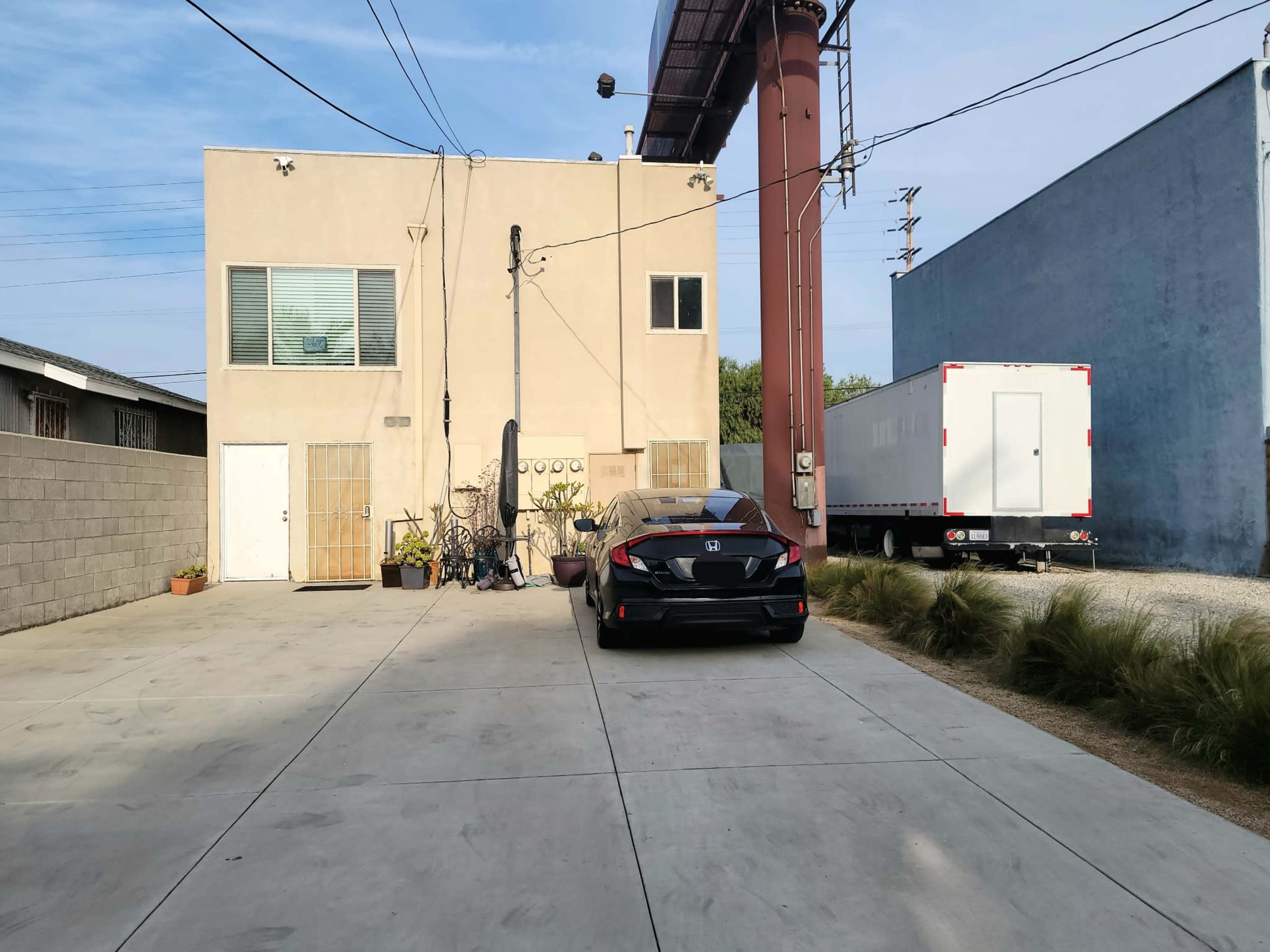 A black car is parked in a concrete lot in front of a two-story building, with another building and a trailer visible in the background.