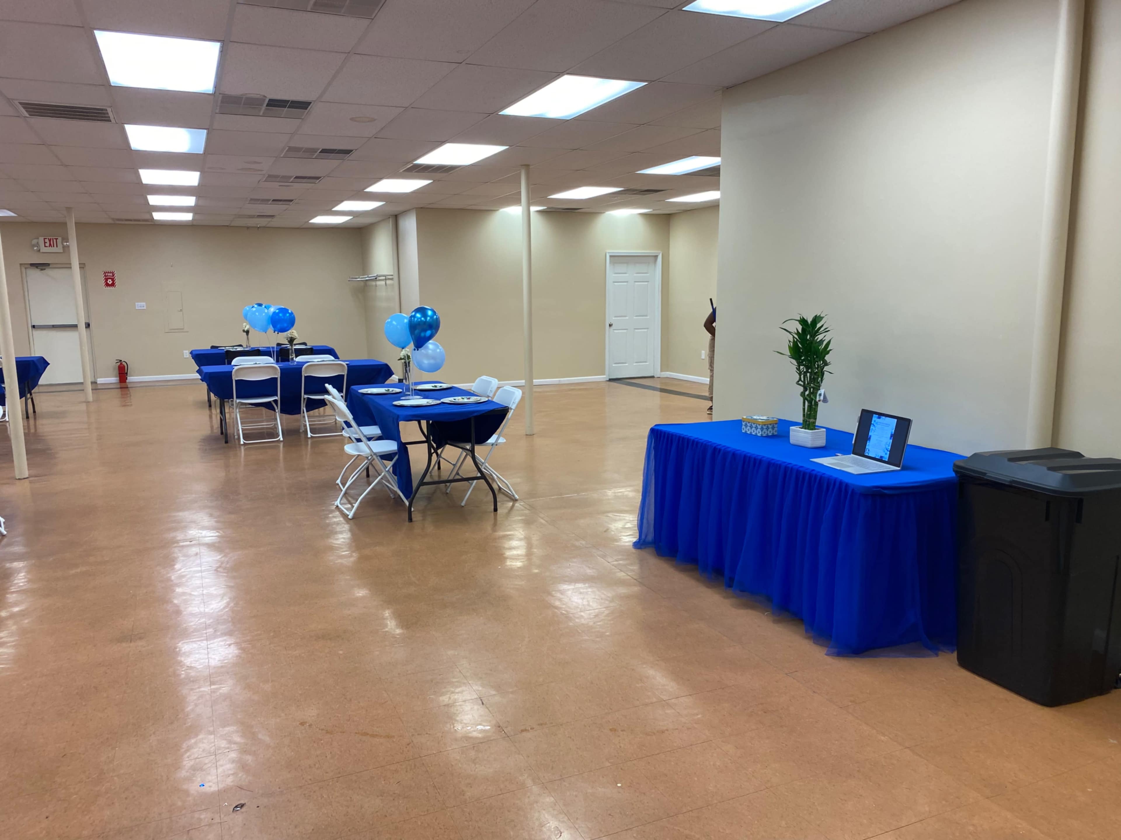 A large, empty room features several tables covered with blue tablecloths and balloons, along with a table displaying decorative items and a trash can.