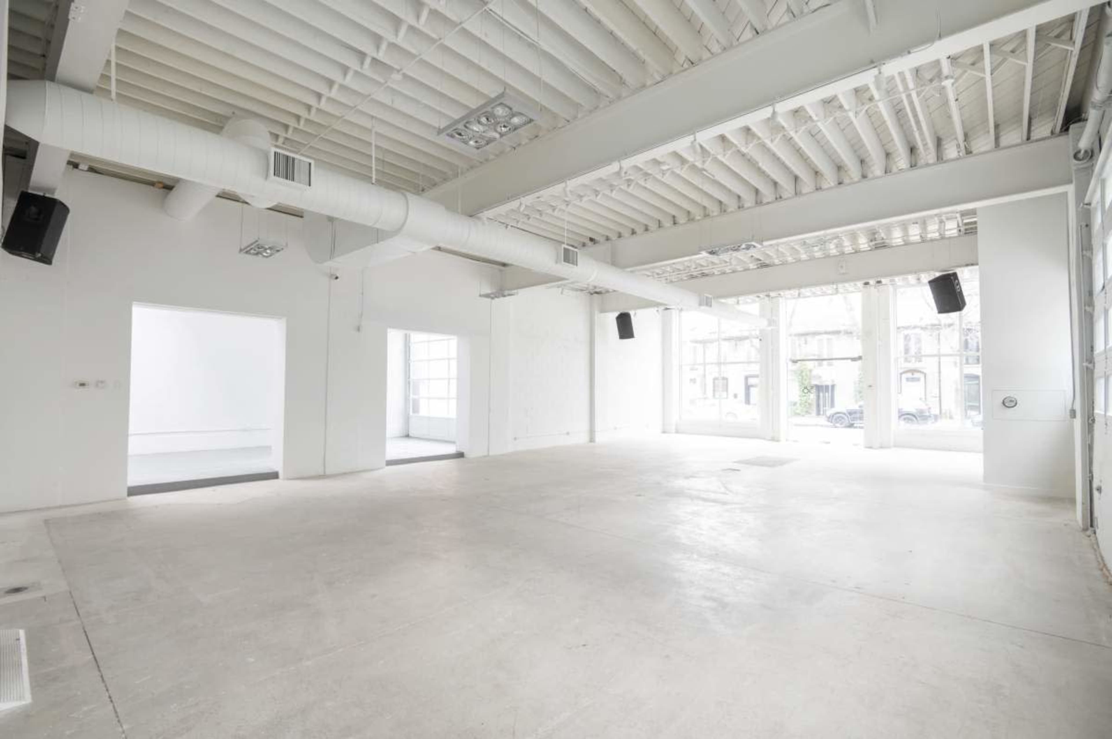 The image shows an empty, well-lit commercial space with white walls and a concrete floor, featuring large windows and high ceilings.