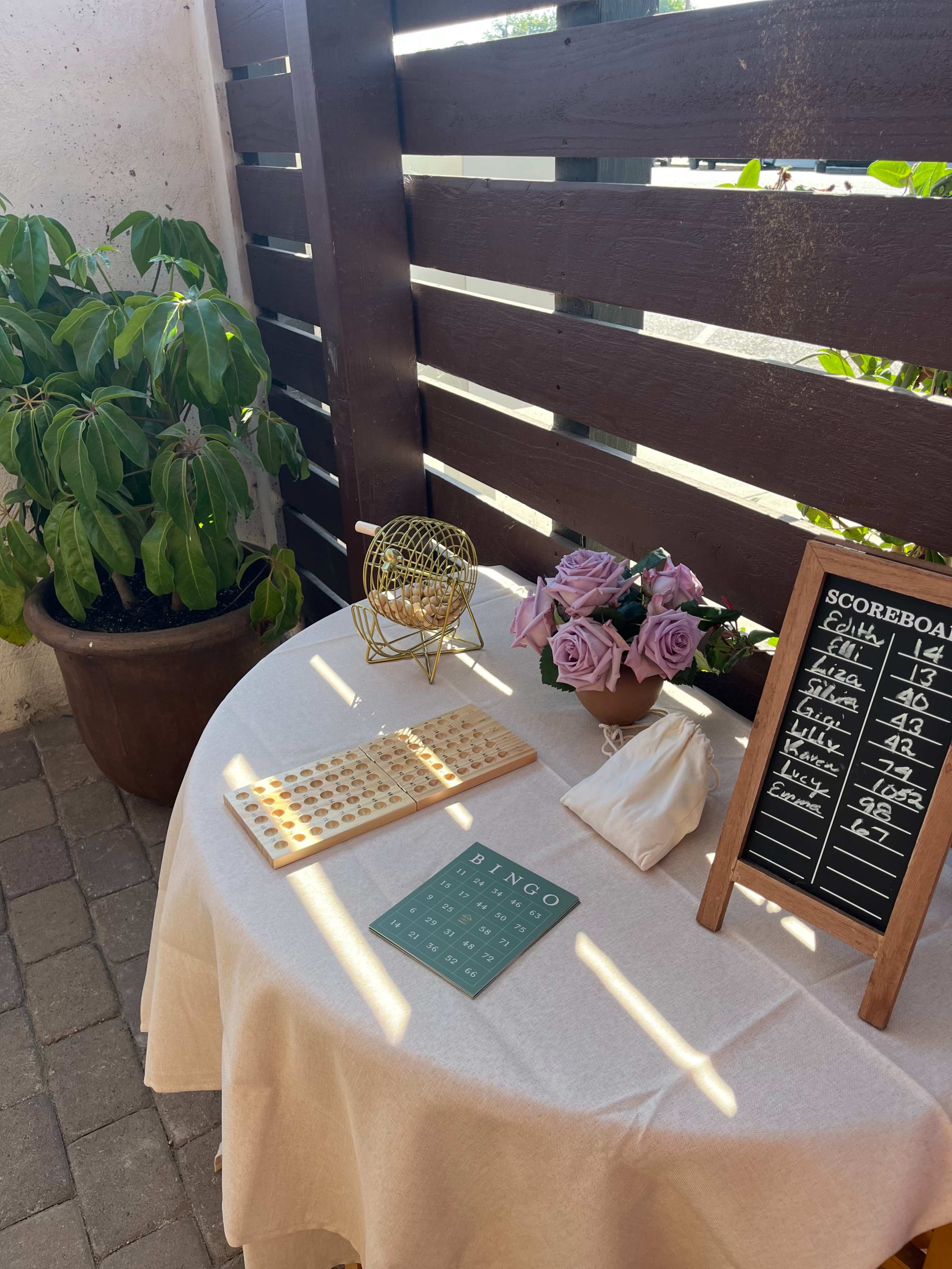 A table is set with a bingo card, wooden markers, a score sheet, and a vase of flowers beside a large indoor plant.