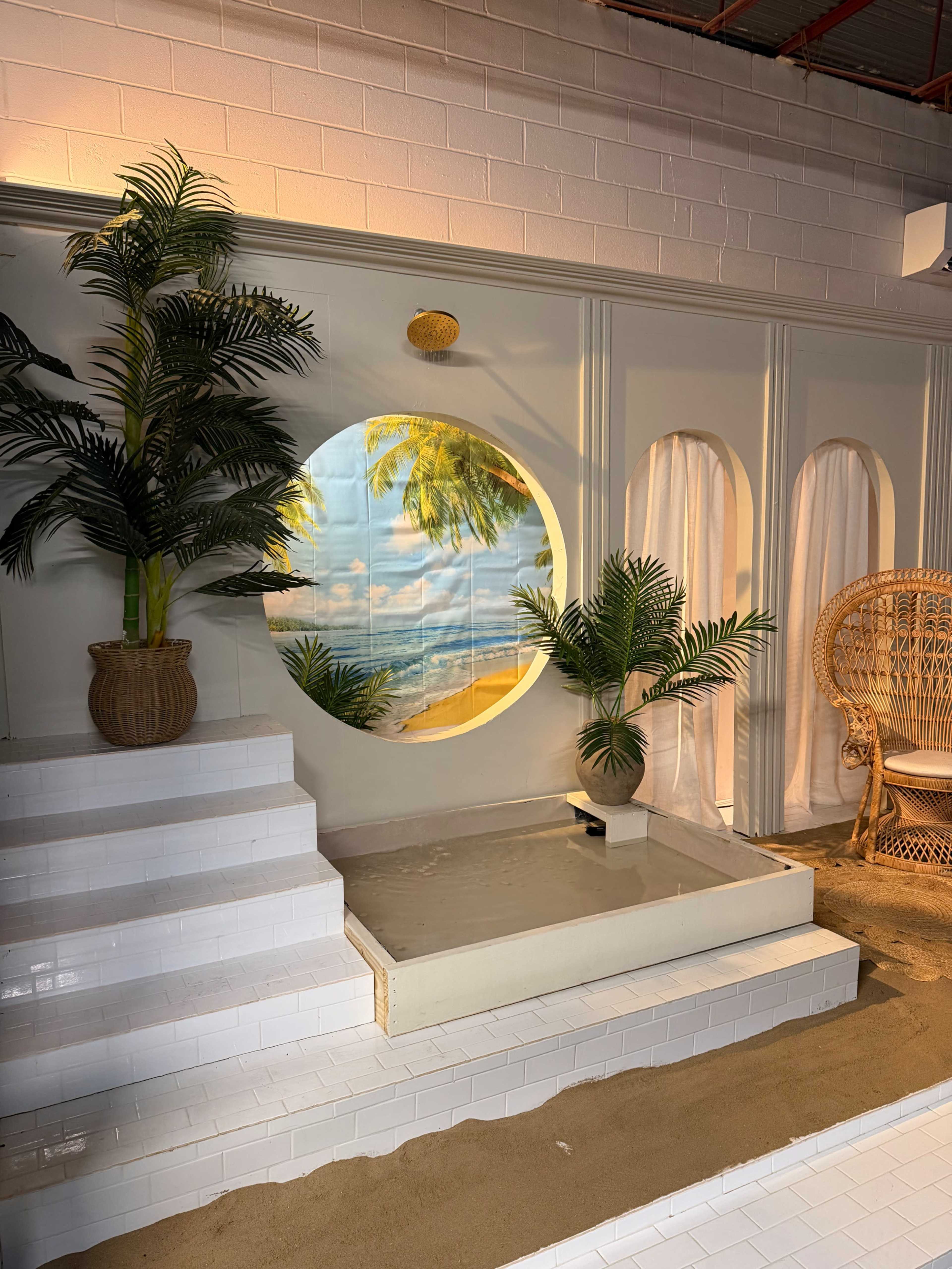 The image depicts a bright indoor setting featuring a circular beach-themed mural, potted plants, and a shallow water feature, along with white tiled steps leading to an area with a wicker chair.