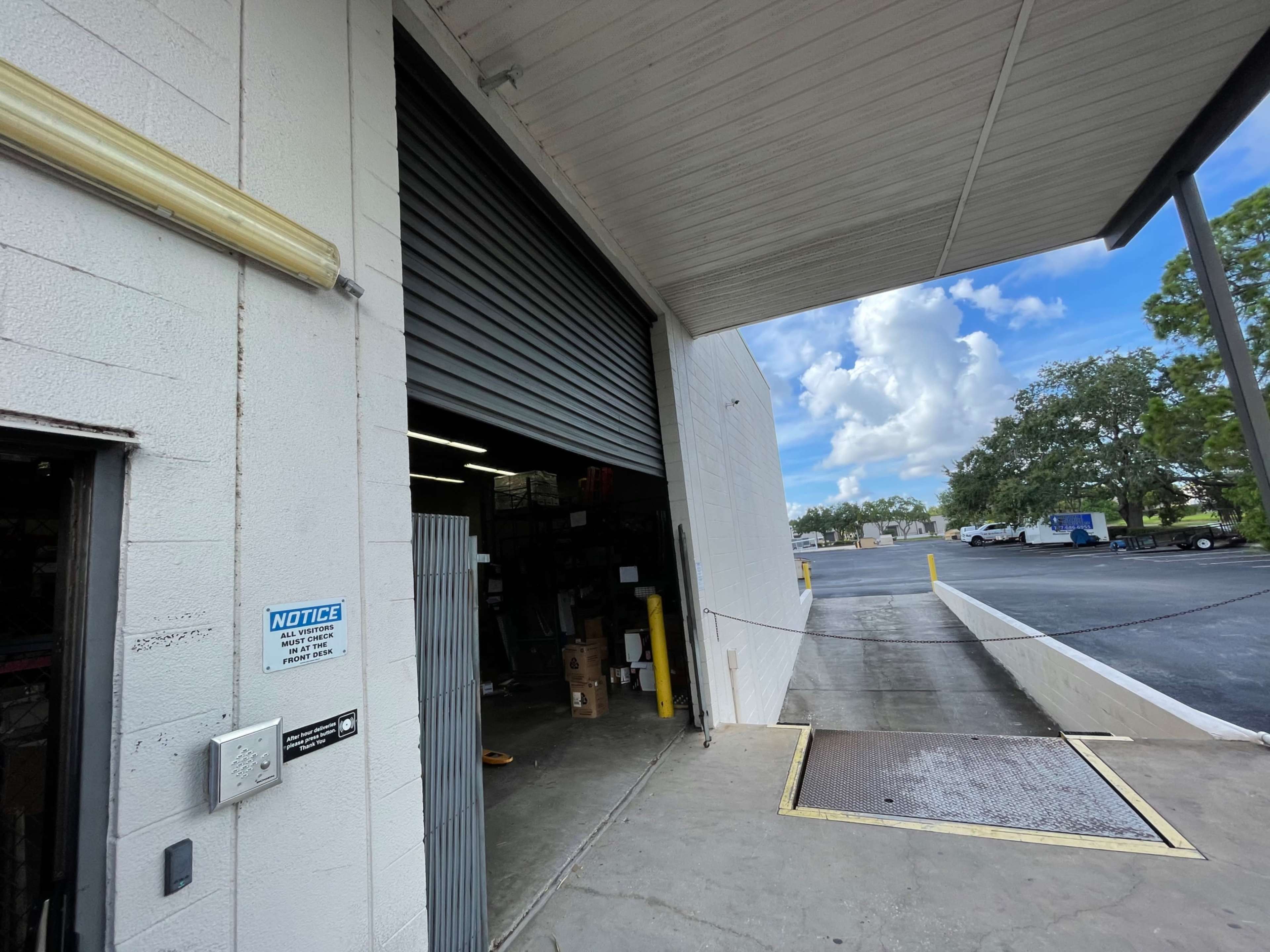 A large warehouse door is partially open, revealing the interior space and a concrete loading area outside beneath a blue sky with scattered clouds.