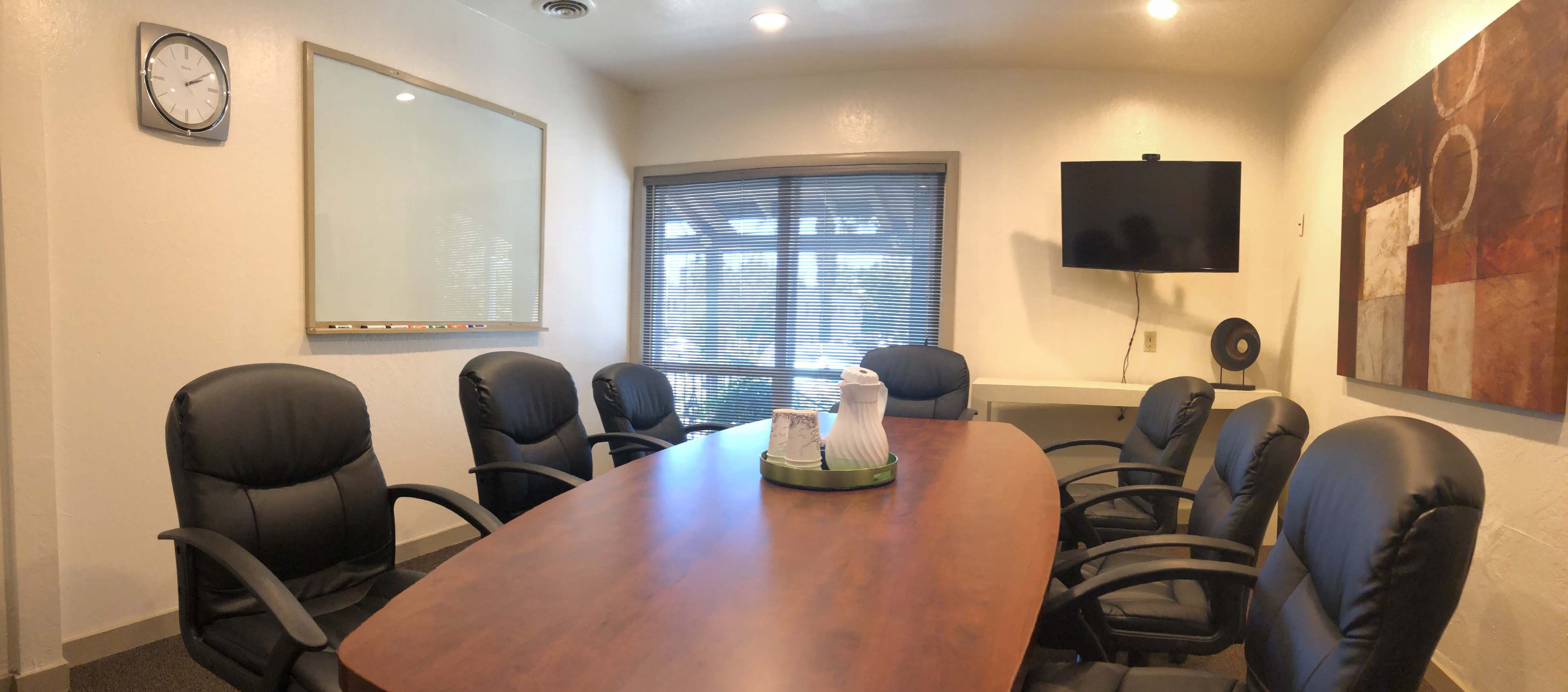 A conference room features a large wooden table surrounded by black ergonomic chairs, a whiteboard, a wall-mounted TV, and a modern art piece.