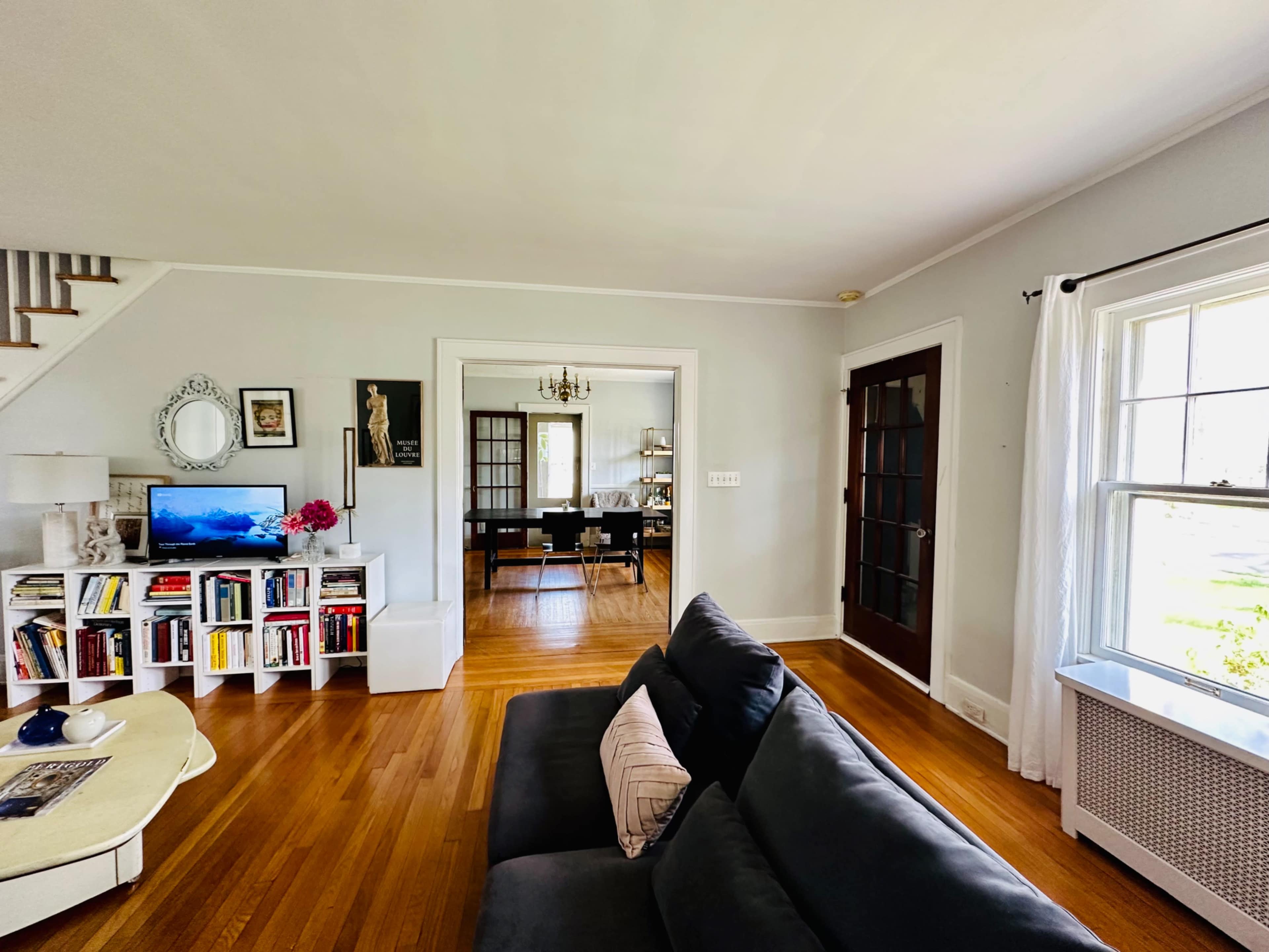A living room features a dark sofa, a bookshelf filled with books, and a doorway leading to a dining area with a chandelier.