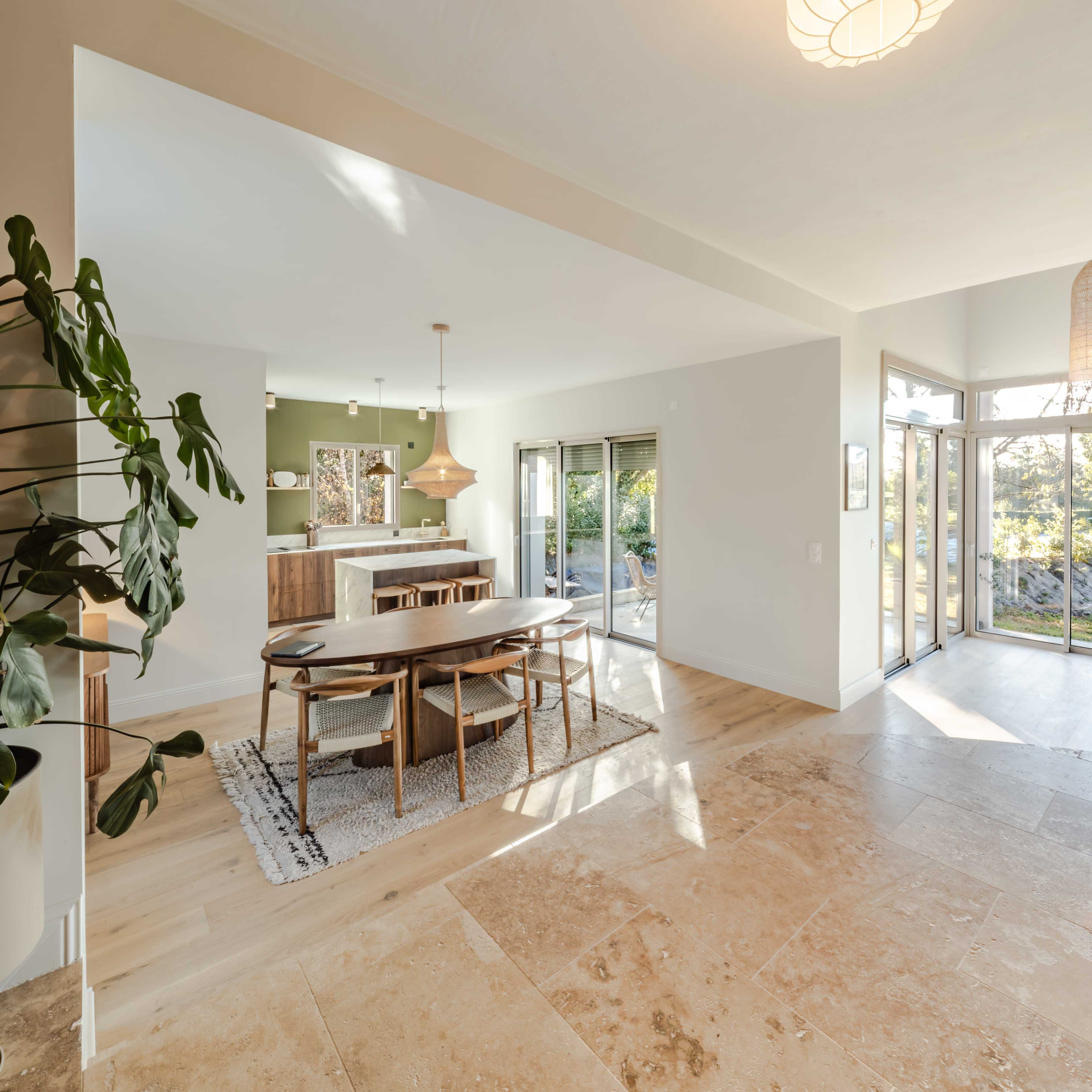 The image shows a spacious, modern dining area with a wooden table and chairs, adjacent to a well-lit kitchen and sliding doors leading to an outdoor area.