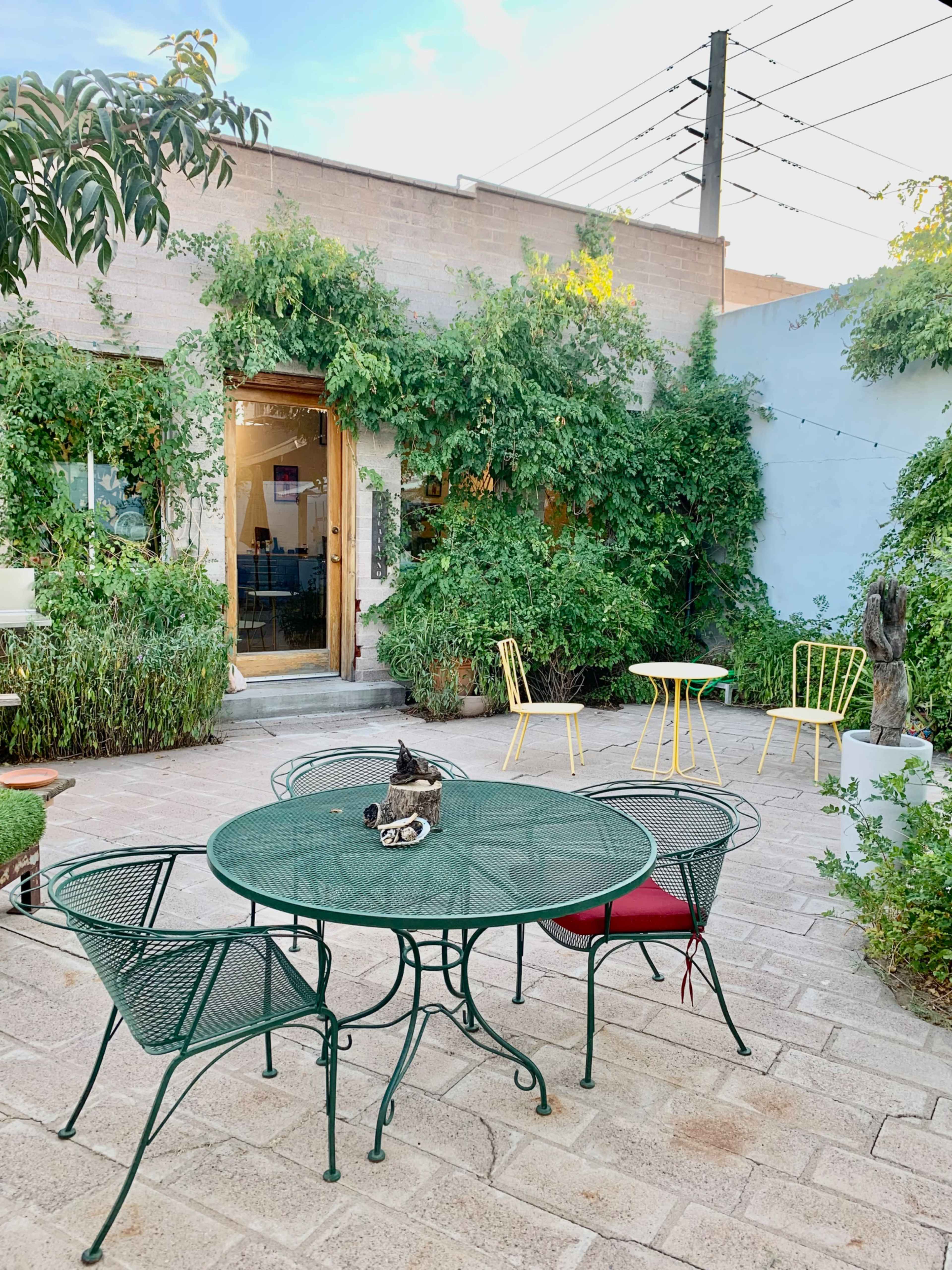 A small outdoor seating area features a green metal table surrounded by four chairs, with lush plants and a building in the background.