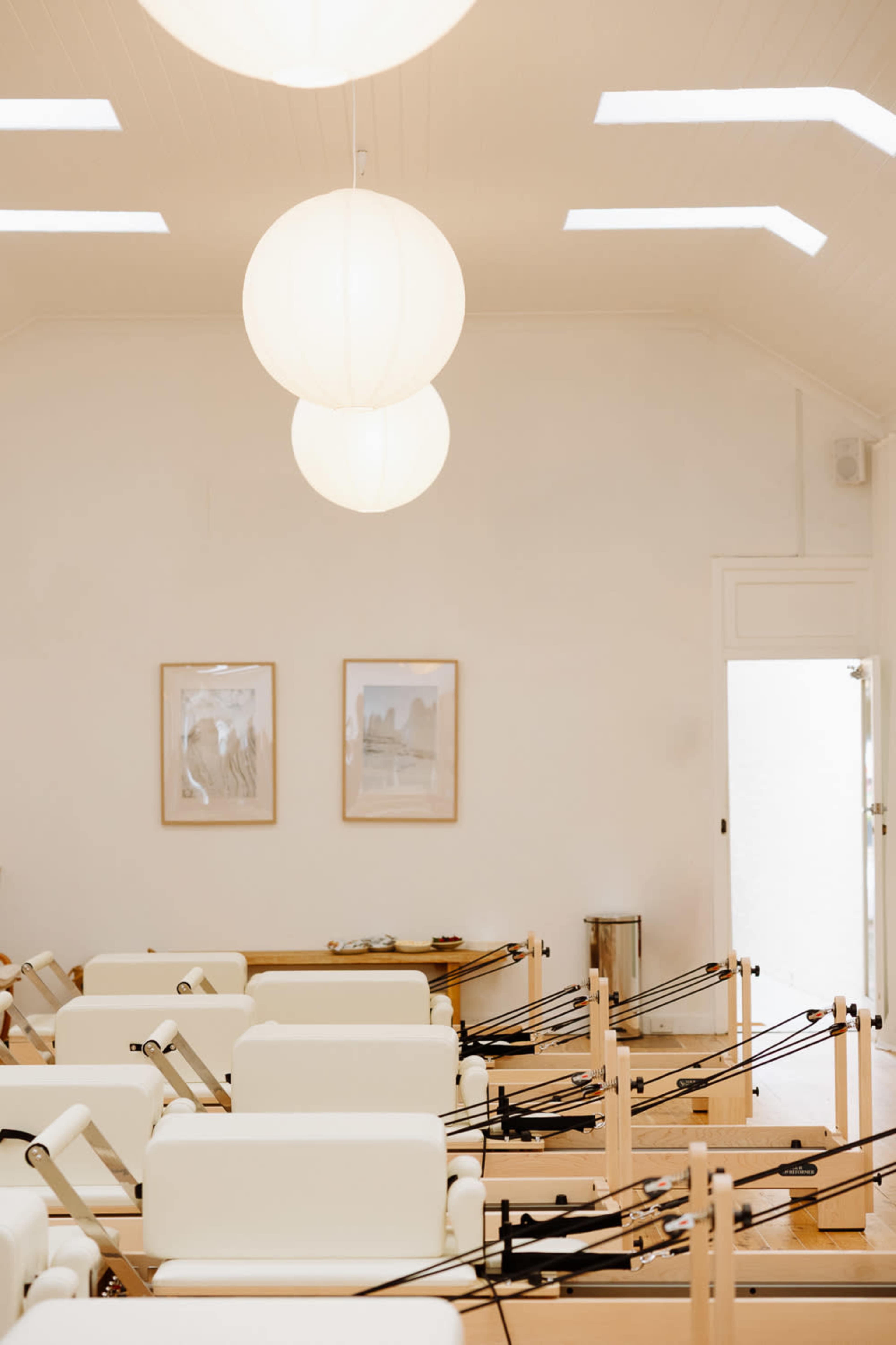 The image shows a bright, minimalist studio with several reformer Pilates machines arranged in a neat layout, illuminated by large round pendant lights.