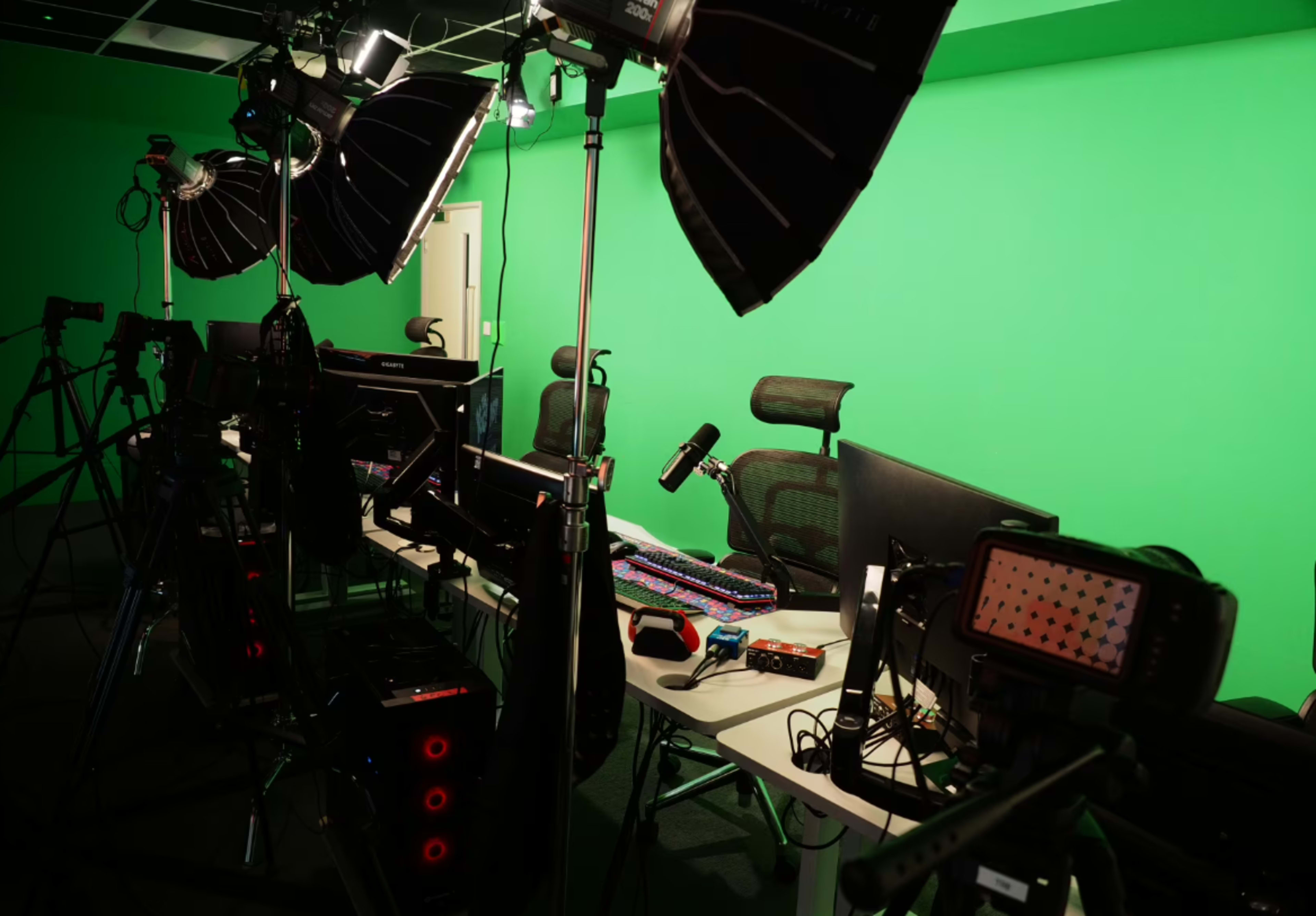 The image shows a studio setup with multiple cameras, lighting equipment, and computers in front of a green screen backdrop.