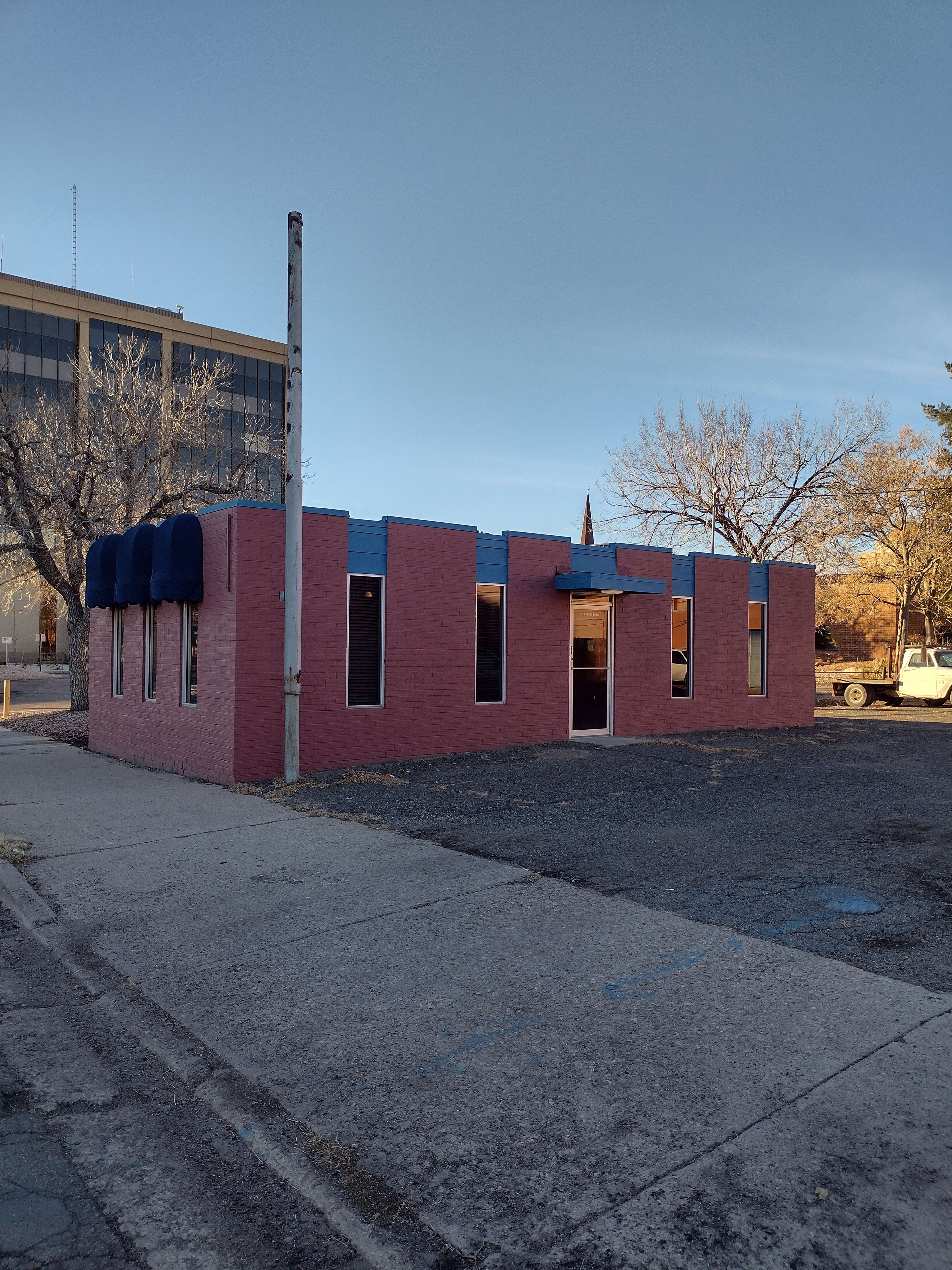 The image shows a small, pink building with blue accents and several windows, situated on a quiet street near a parking lot.