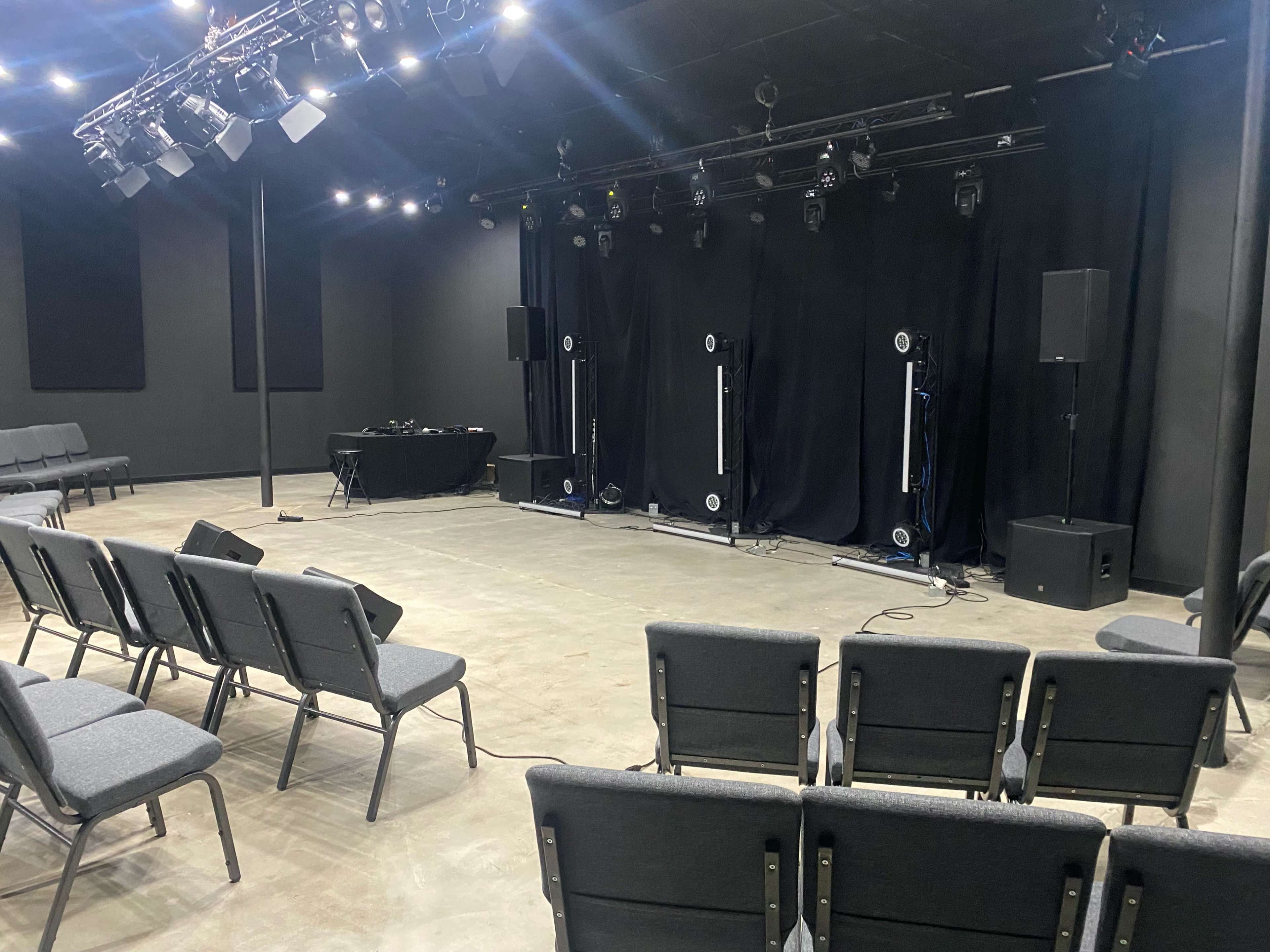 A performance space features rows of gray chairs facing a stage with black curtains and sound equipment.