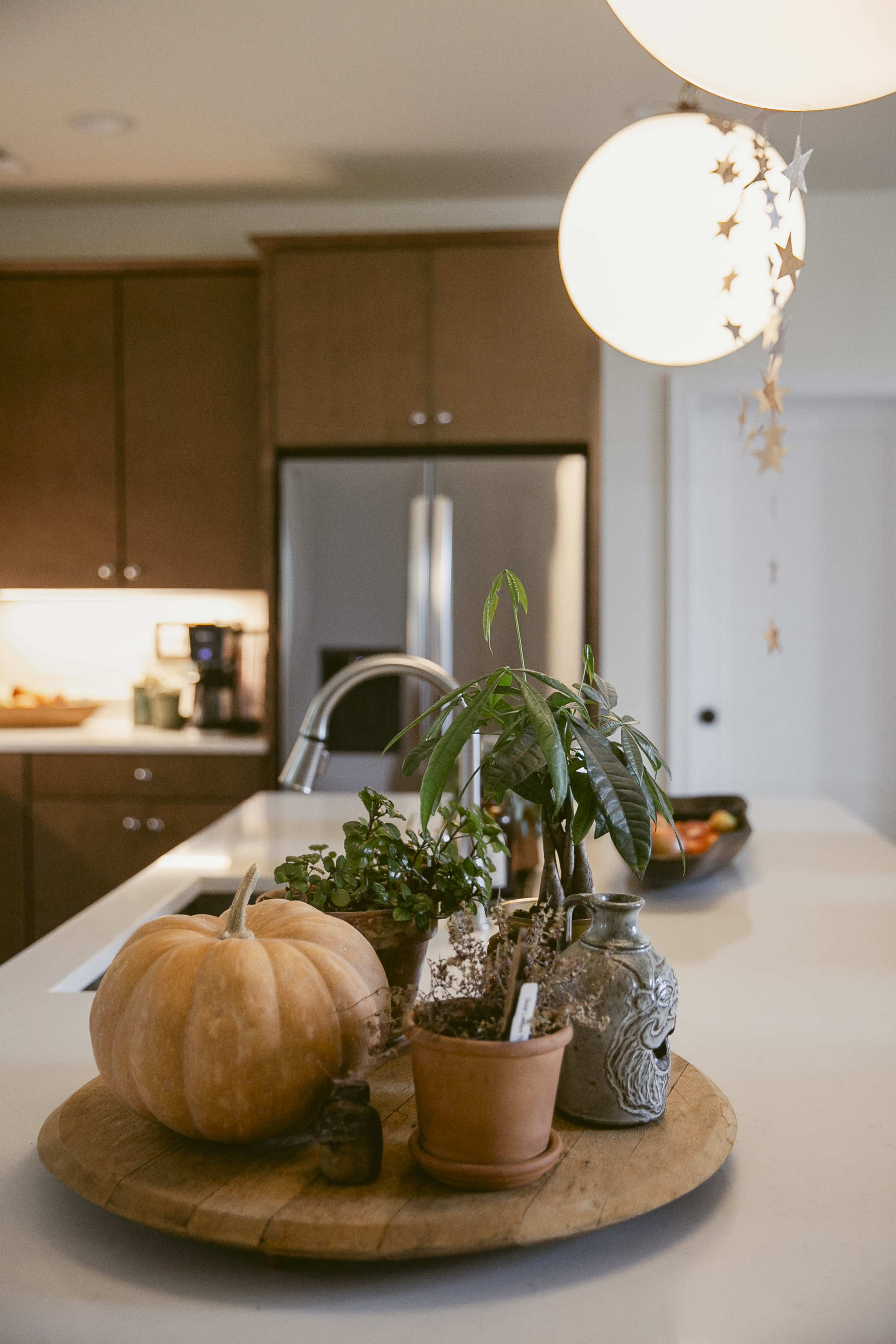 A wooden platter on a kitchen countertop holds a pumpkin and potted plants, with modern pendant lighting hanging above.