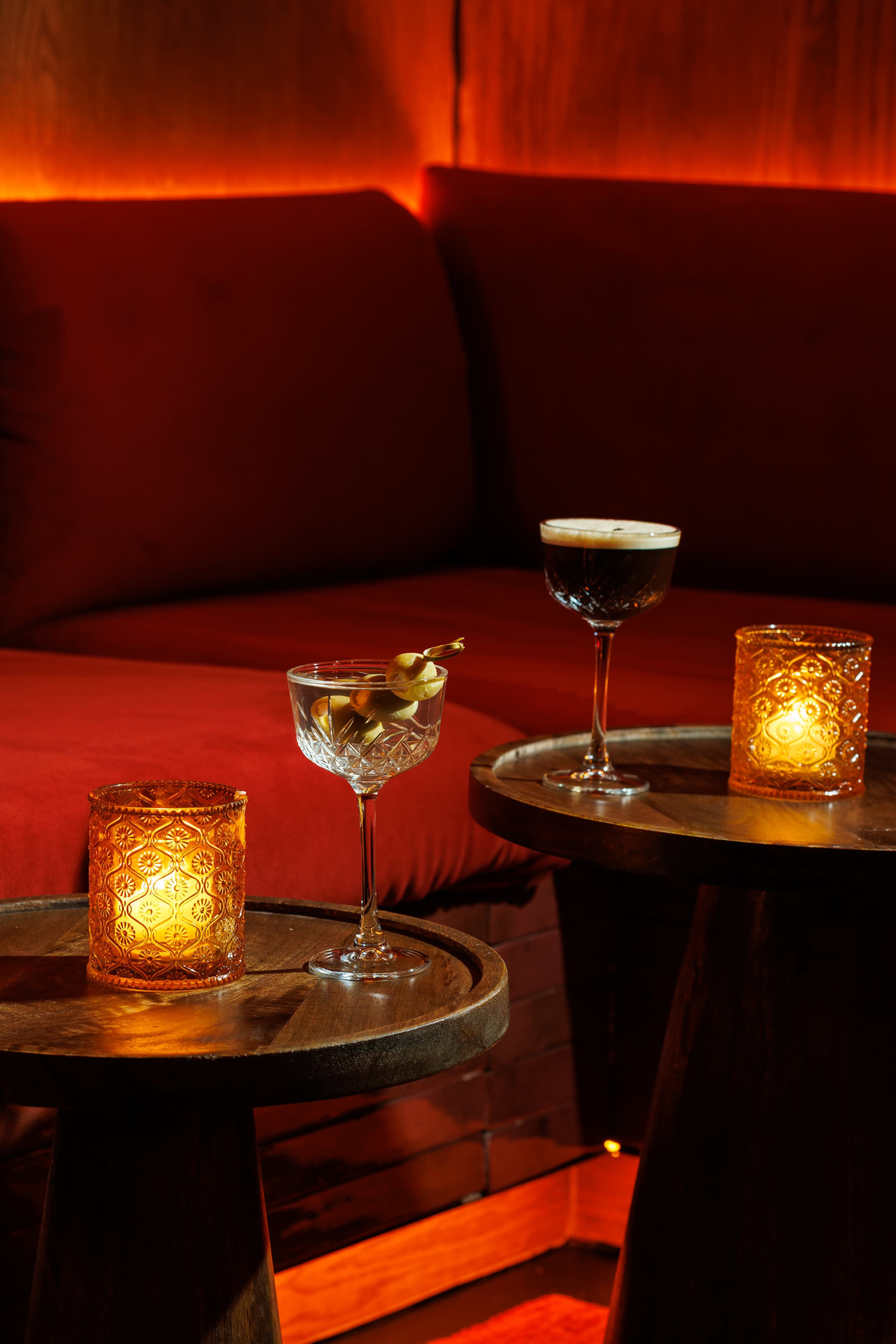 The image shows two cocktails and lit candles on wooden side tables in a dimly lit lounge area with red seating.