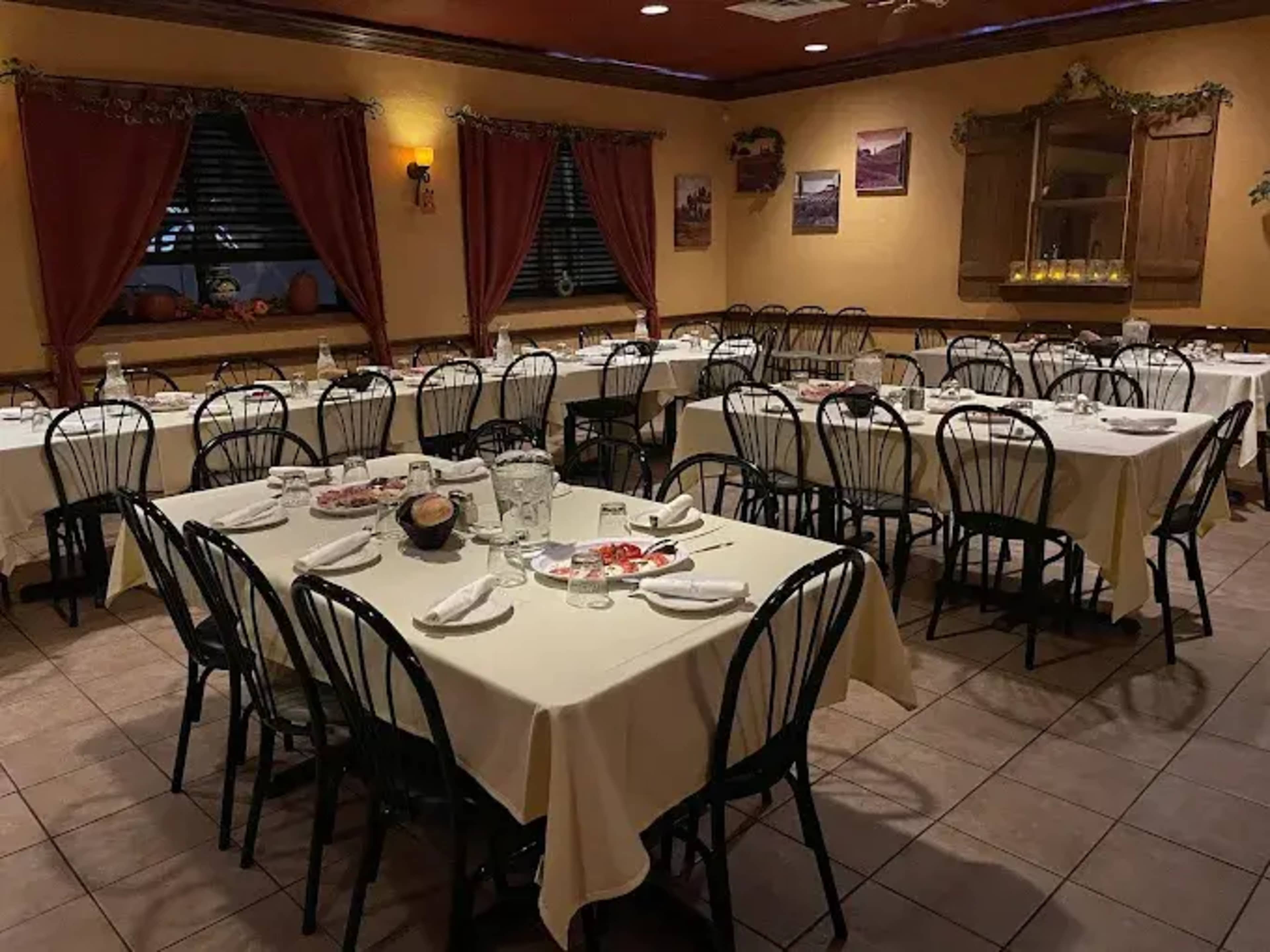 The image shows a dining room with several tables covered in white tablecloths, arranged with black chairs and decorative curtains at the windows.