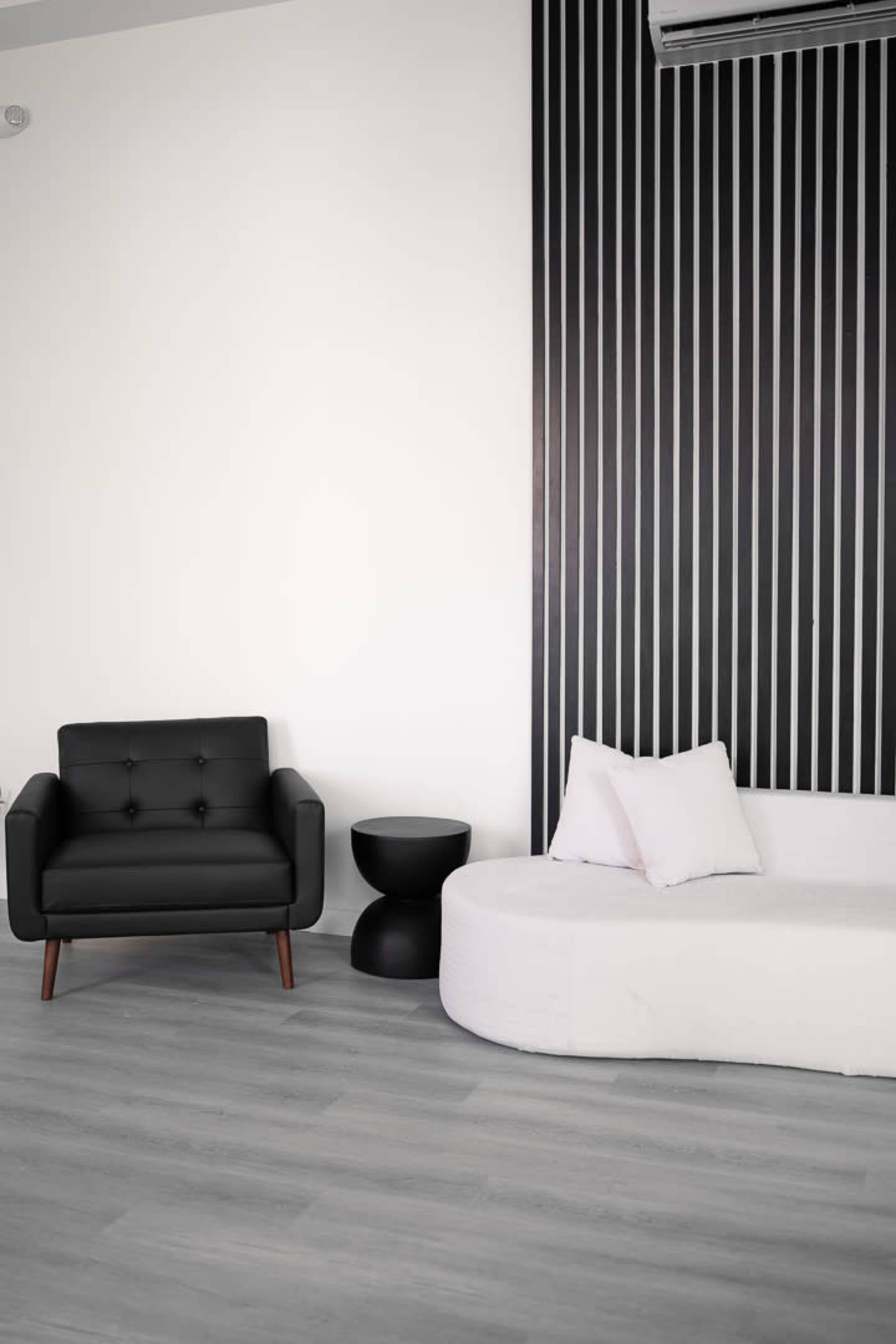 A black armchair and a white, curved sofa are positioned against a striped wall in a minimalist room with light wooden flooring.