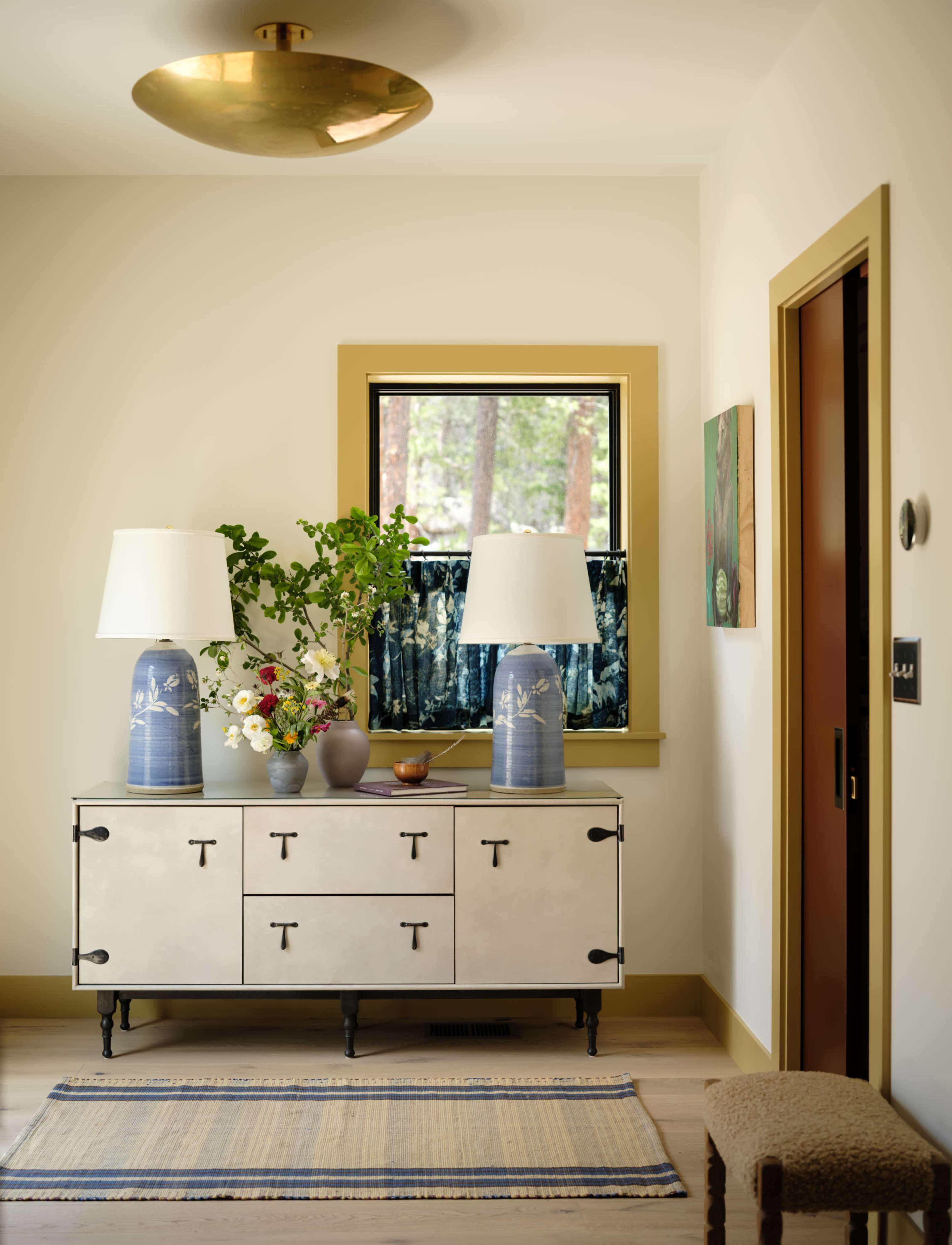 A stylish entryway features a light-colored console table with two blue lamps, a floral arrangement, and a window displaying a view of the outdoors.