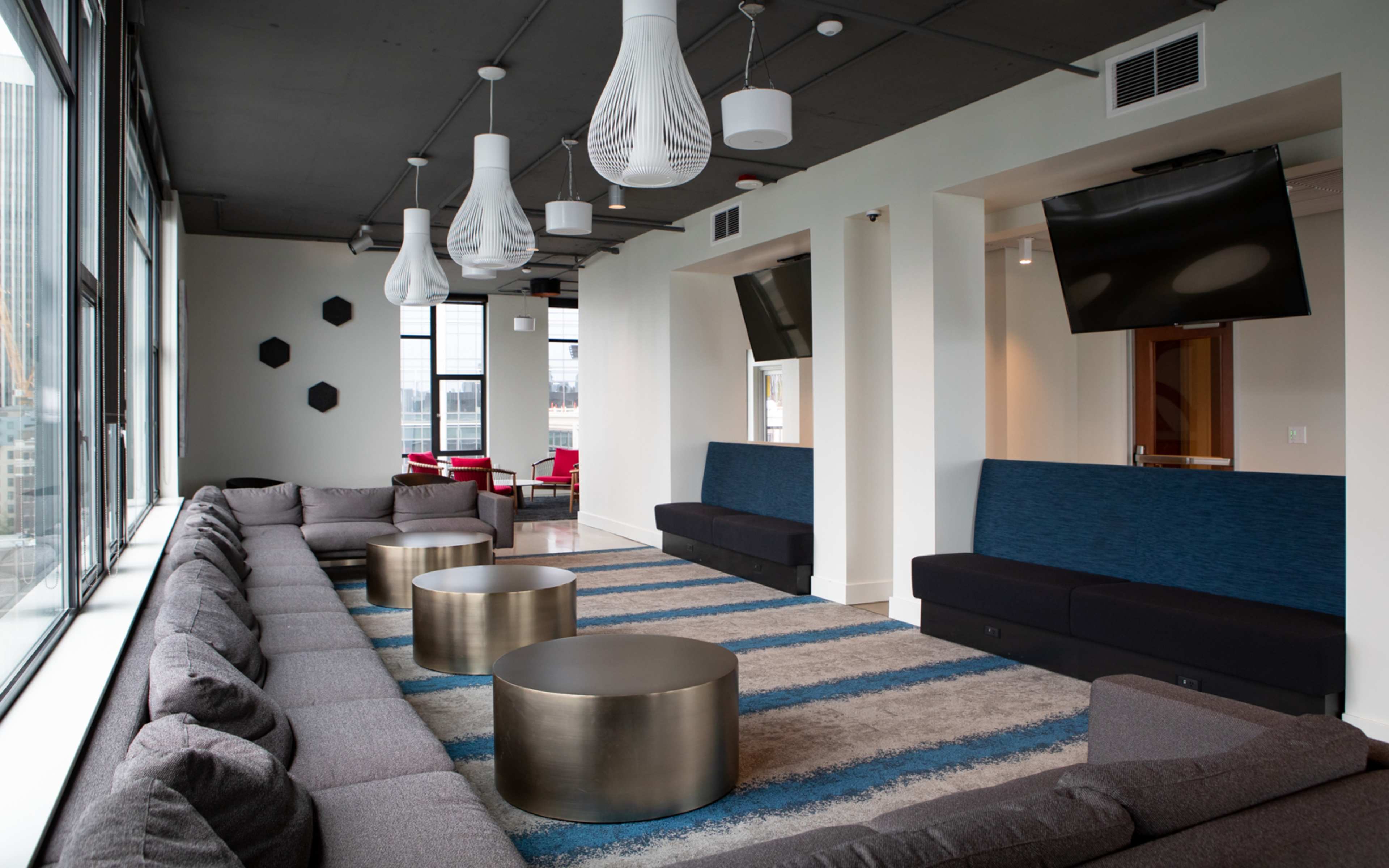 The image shows a modern lounge area with a large, sectional couch, two round metal tables, and unique pendant lights, all situated by large windows.