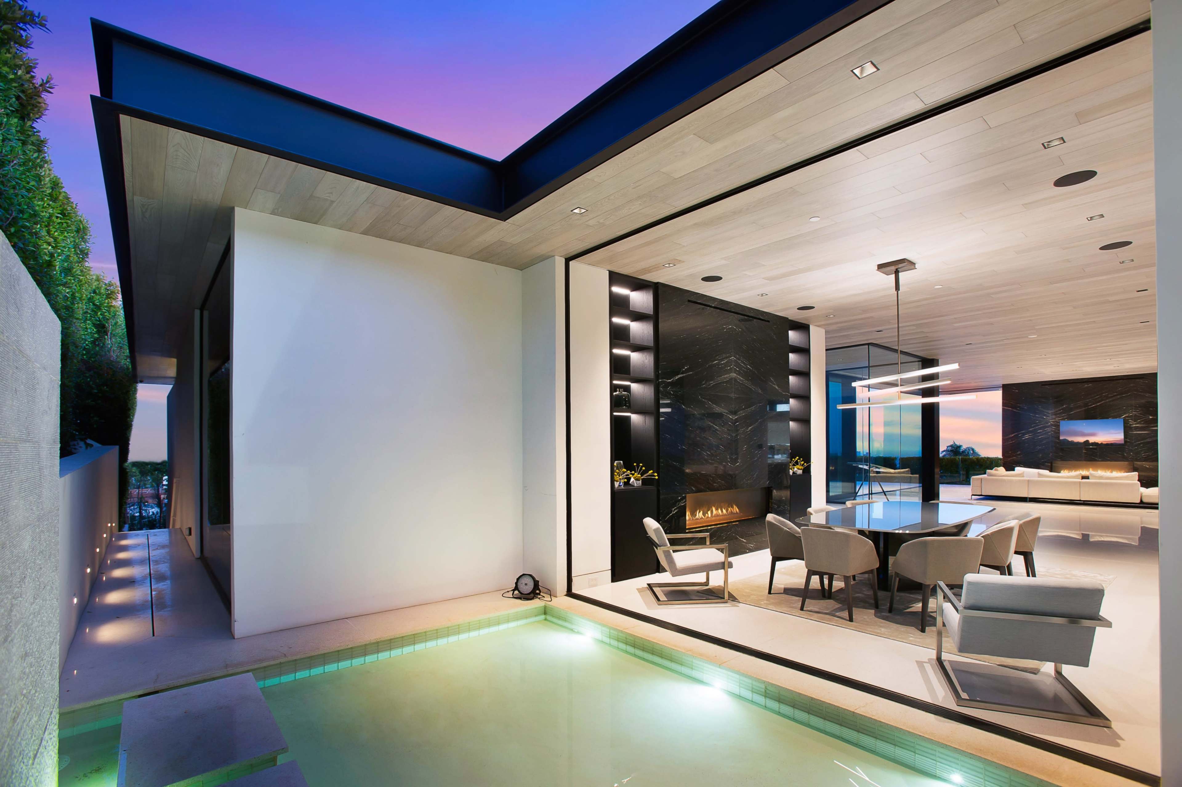 A modern home features a spacious living area with large windows overlooking a pool and outdoor seating area, illuminated by soft evening light.