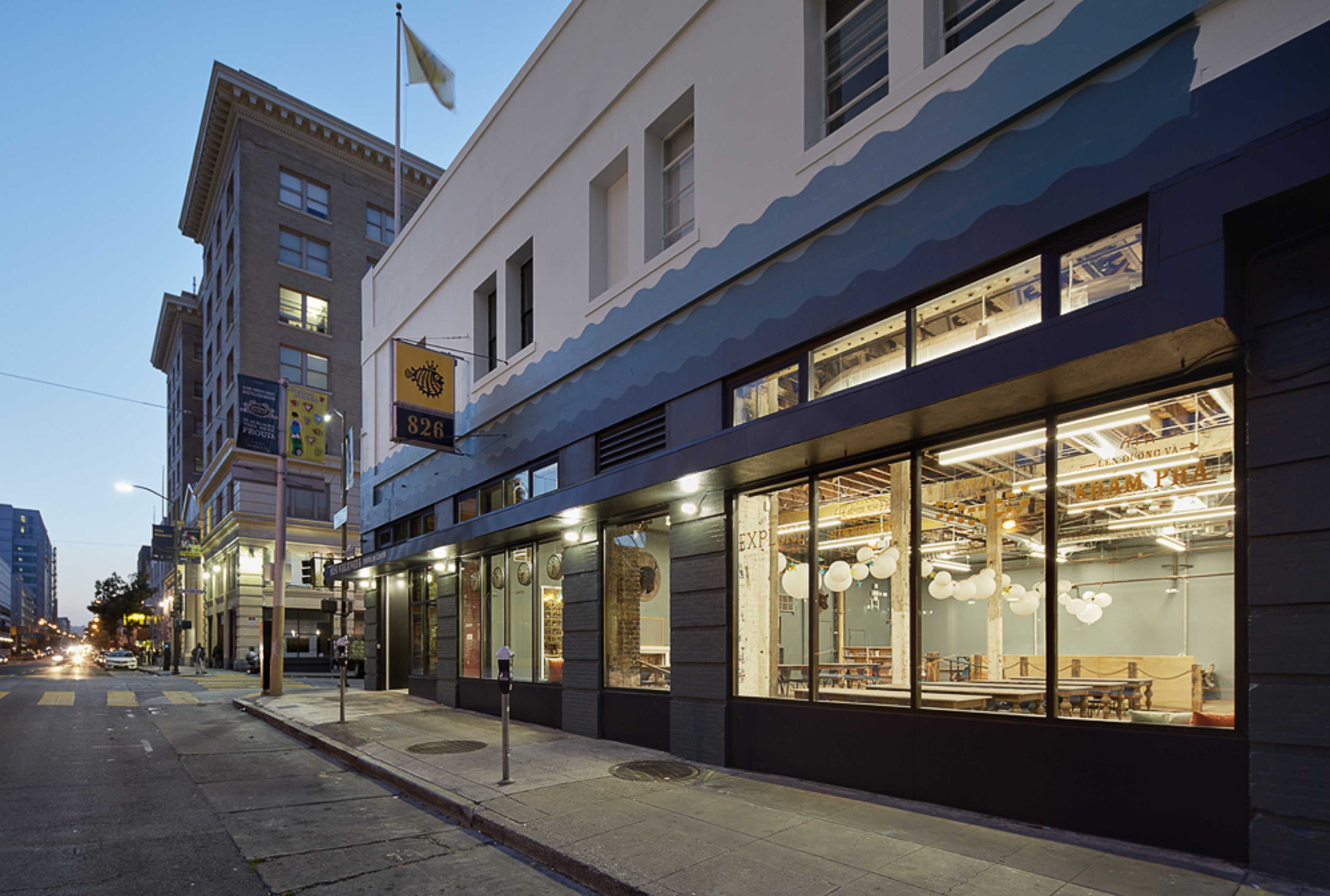 A modern restaurant with large windows, located on a street corner in an urban area, during twilight.