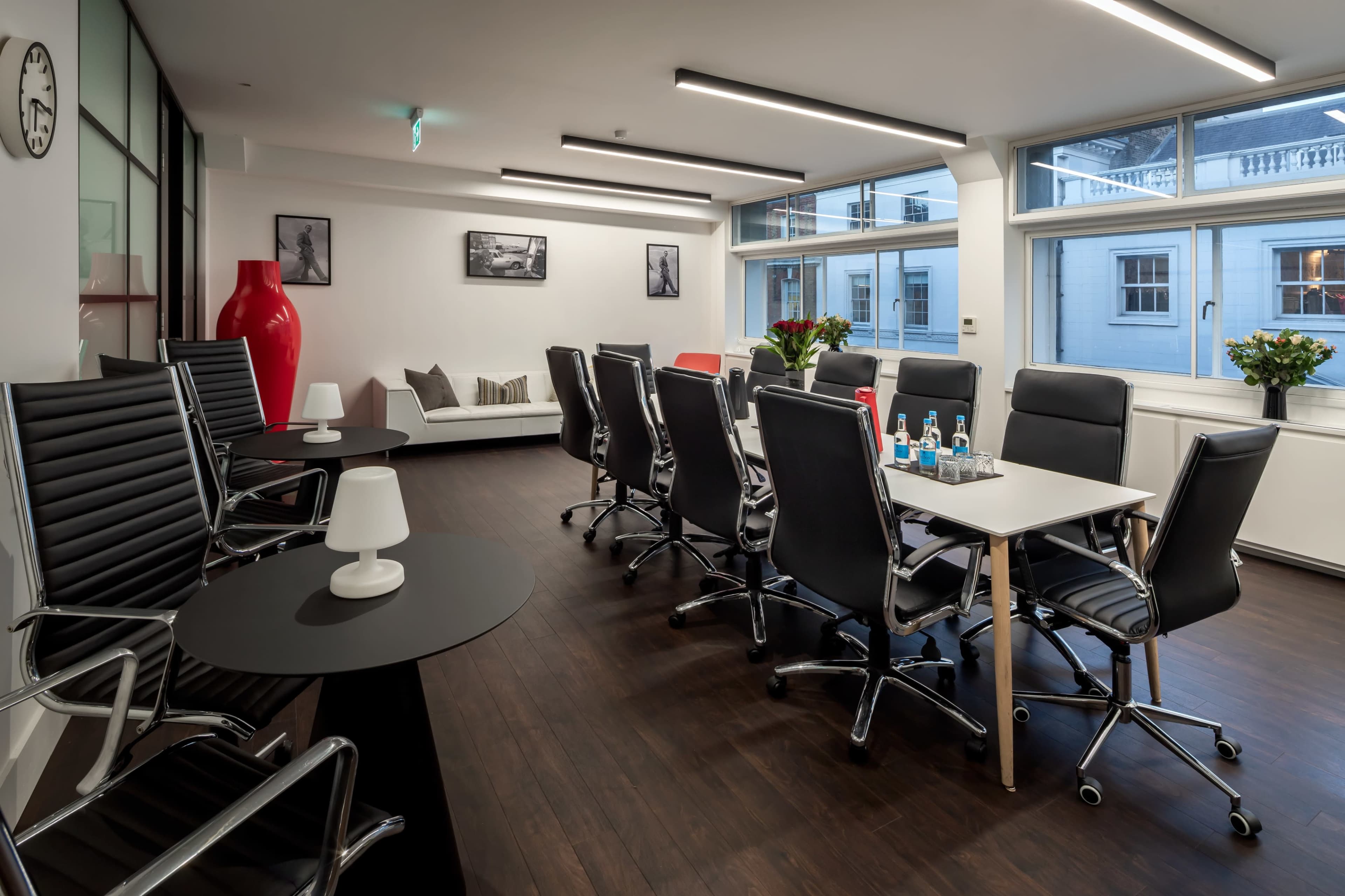 A modern conference room with a long table surrounded by black and red chairs, large windows providing natural light, and a few decorative elements like vases and artwork.