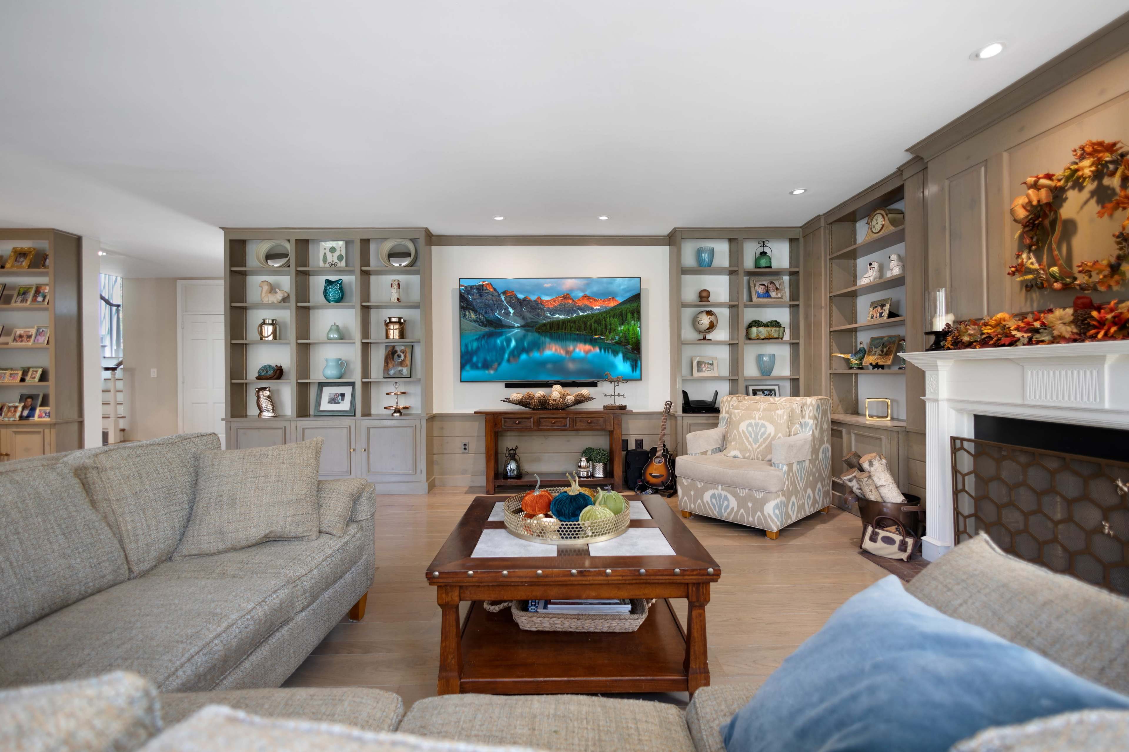 A modern living room features a sectional sofa, a coffee table, shelves with decorative items, and a mounted television displaying a scenic landscape.