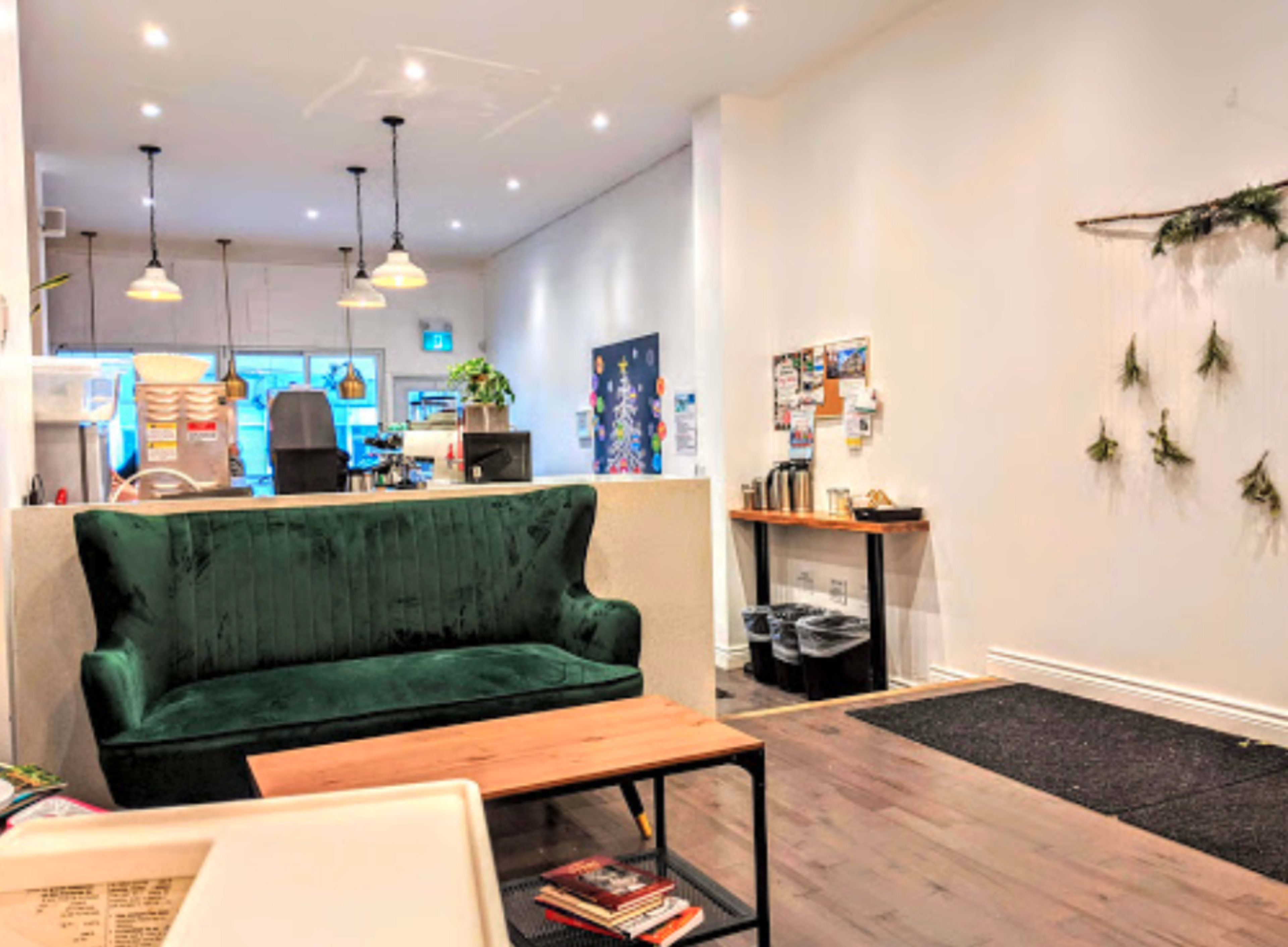 The image shows a cozy café interior featuring a green velvet sofa, wooden coffee table, and a wall adorned with plants and bulletin boards.