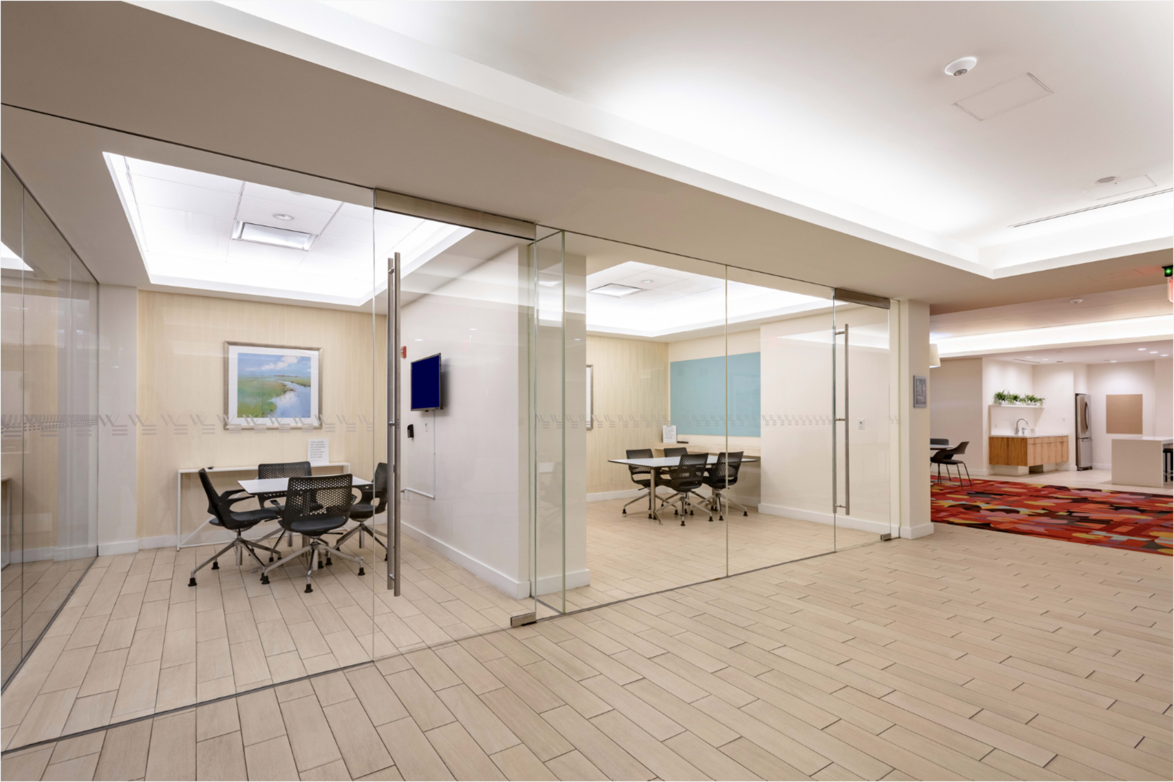 The image shows a modern office space featuring glass walls, a conference area with tables and chairs, and light-colored decor.