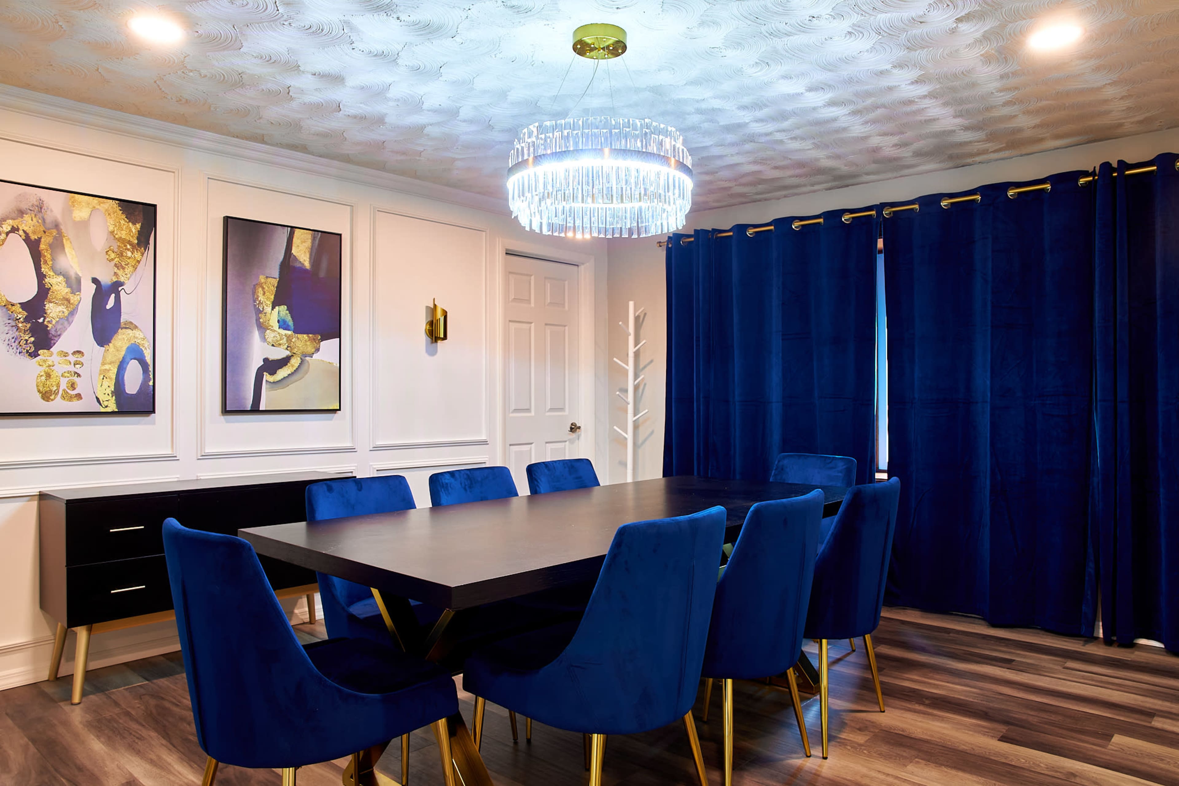 A modern dining room features a long black table surrounded by blue velvet chairs, with a chandelier overhead and dark curtains framing the window.